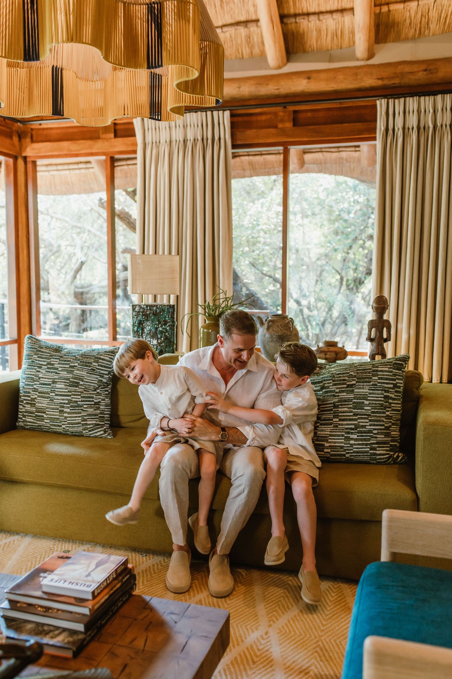 Father sitting on a green sofa with two children in a warm African safari lodge living room.