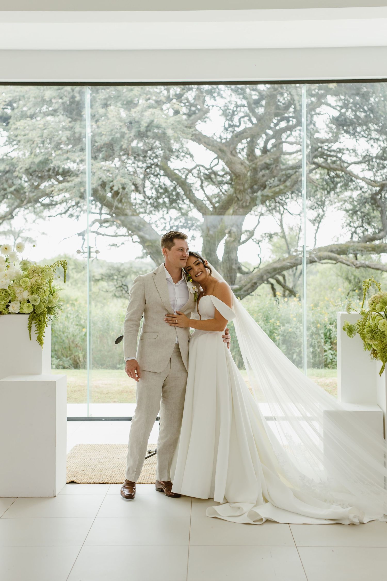 Wedding couple sharing intimate moment in sleek white venue with floor-to-ceiling windows.
