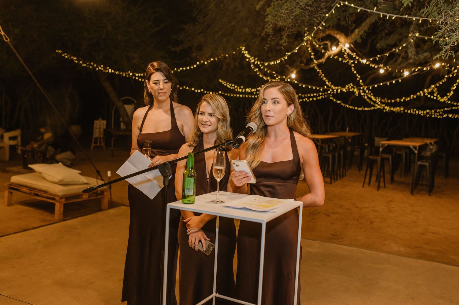Three attendants in dark dresses give speeches at an outdoor evening event.