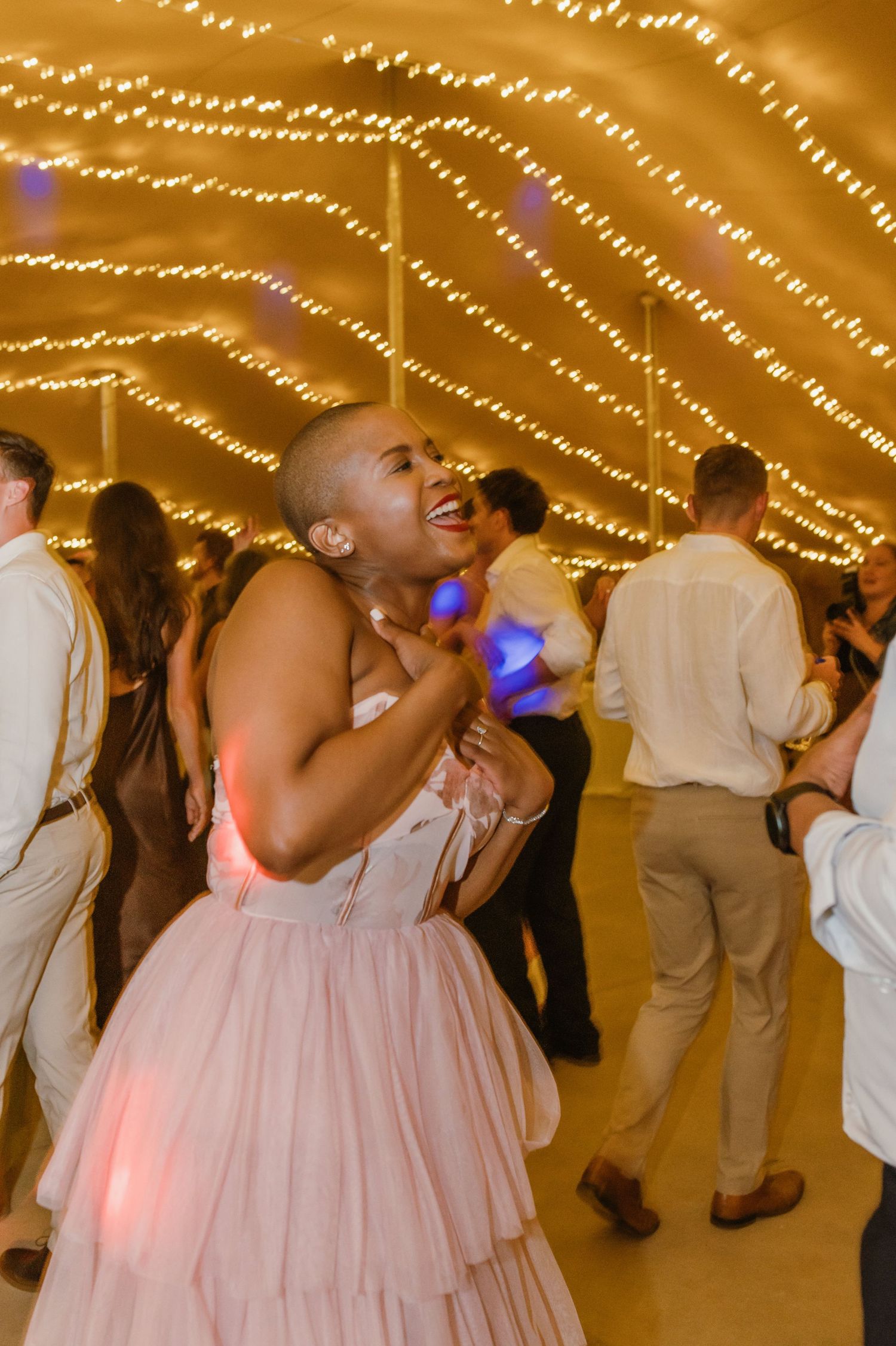 Wedding guests in formal attire dancing enthusiastically under string lights at an evening reception.