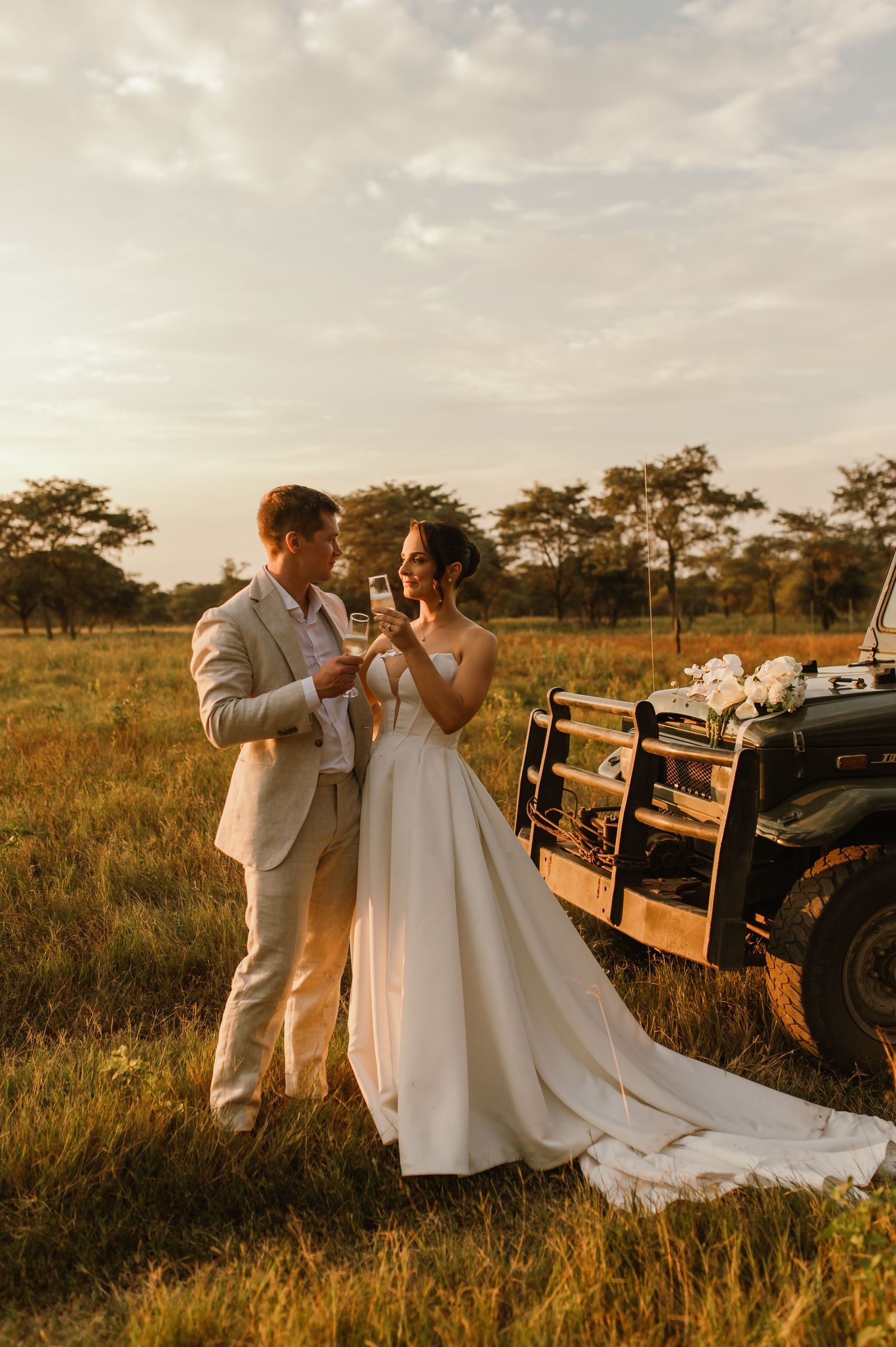 A couple shares champagne during their sunset wedding photoshoot near a vintage vehicle.