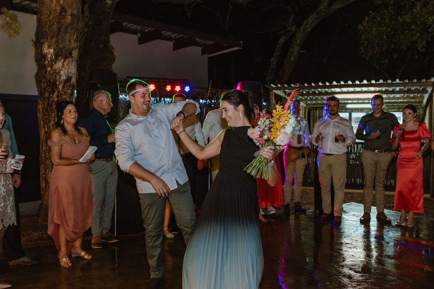 Wedding guest catches bridal bouquet toss outdoors at night reception with crowd watching and cheering.