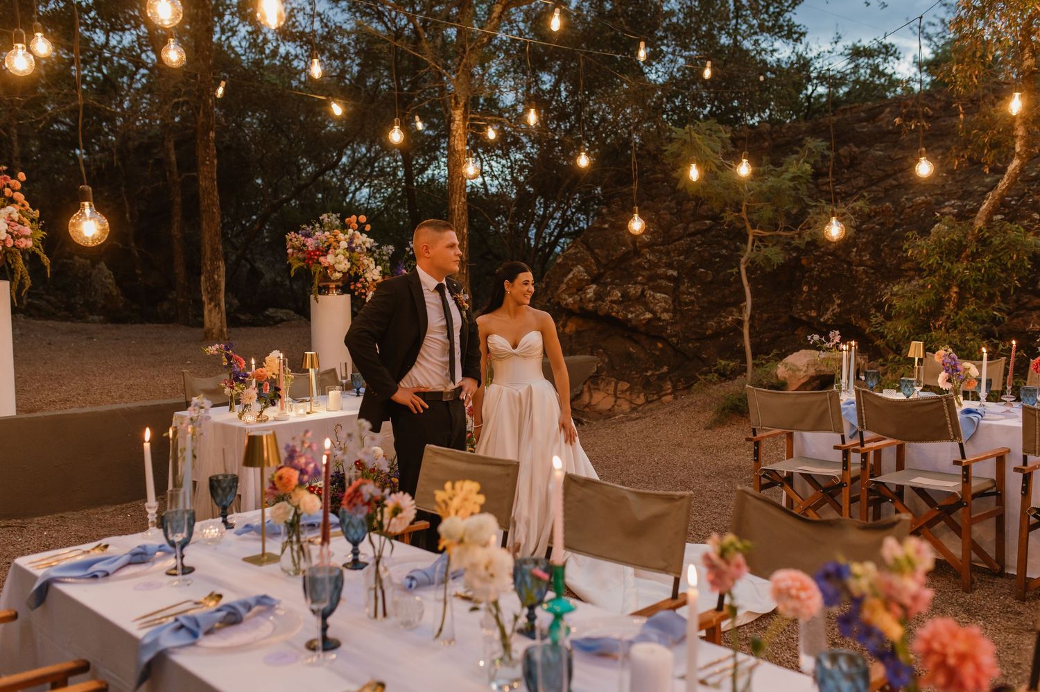 Bride and groom stand at elegantly decorated outdoor reception table with string lights at dusk.