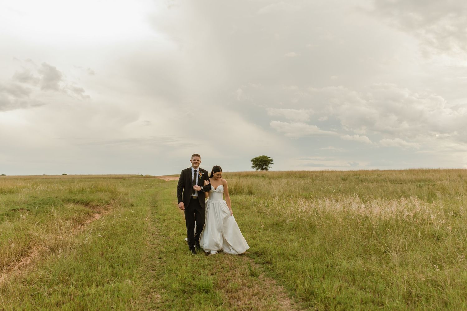 Bride and groom walking hand in hand through open savanna field after wedding.