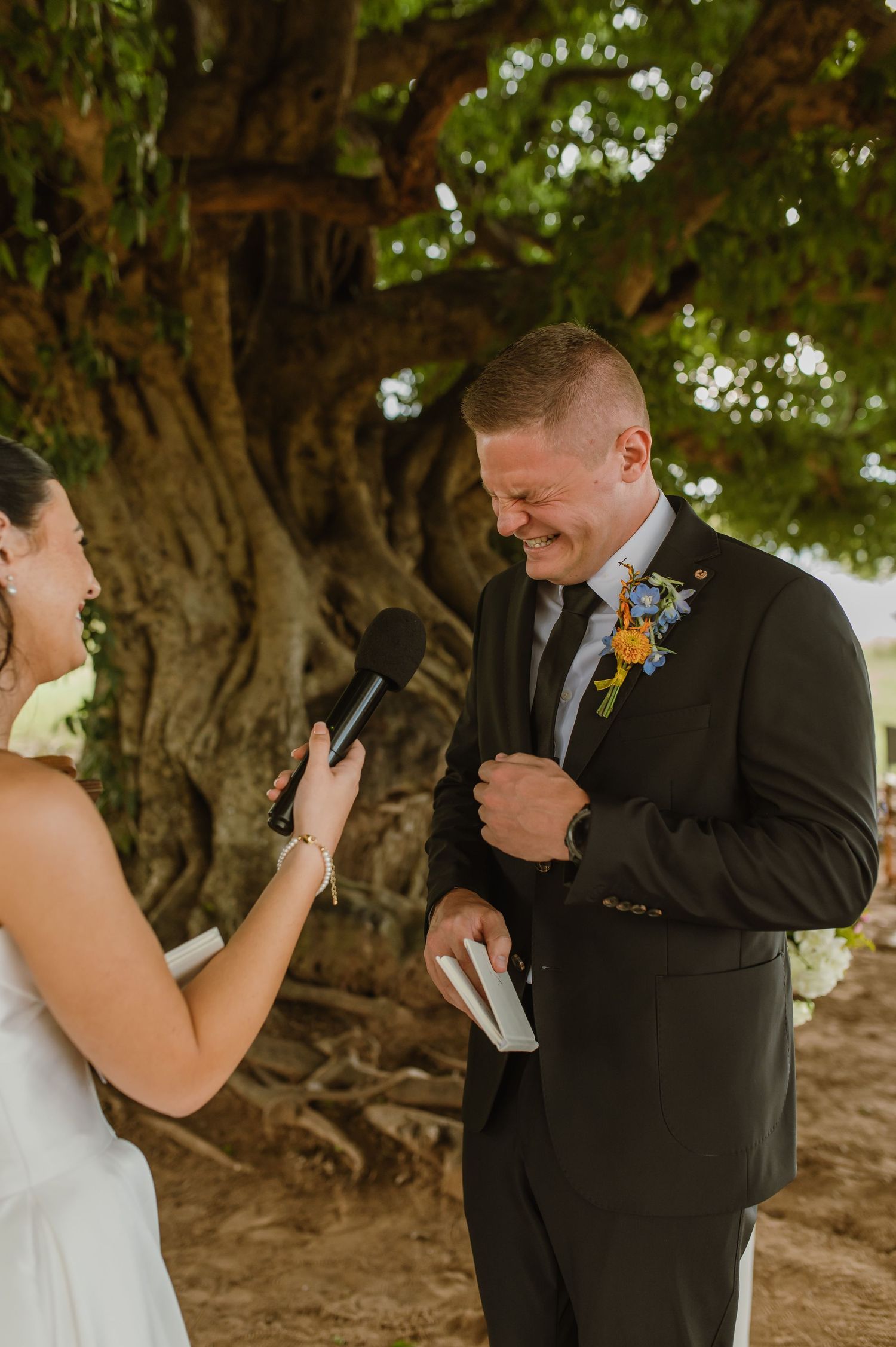 Groom laughing joyfully during wedding vows under a large tree, wearing a dark suit with colorful boutonniere.