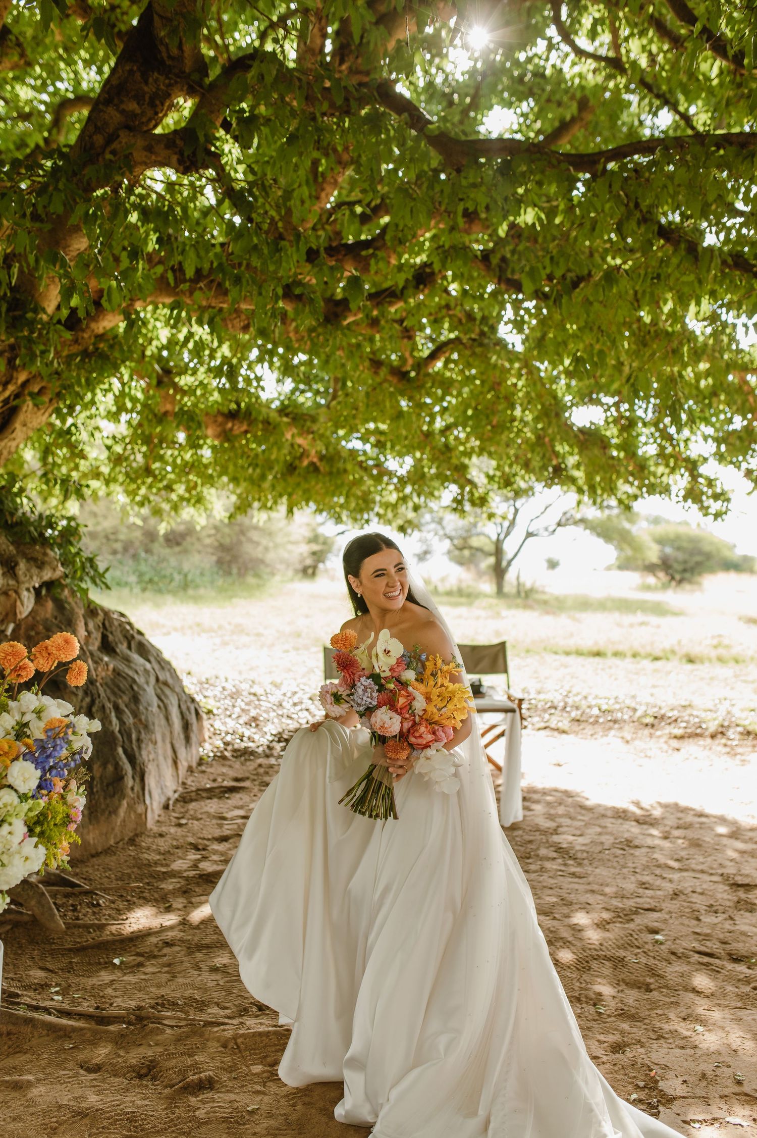 Joyful bride in white gown holding colorful bouquet standing under sunlit tree in outdoor setting.