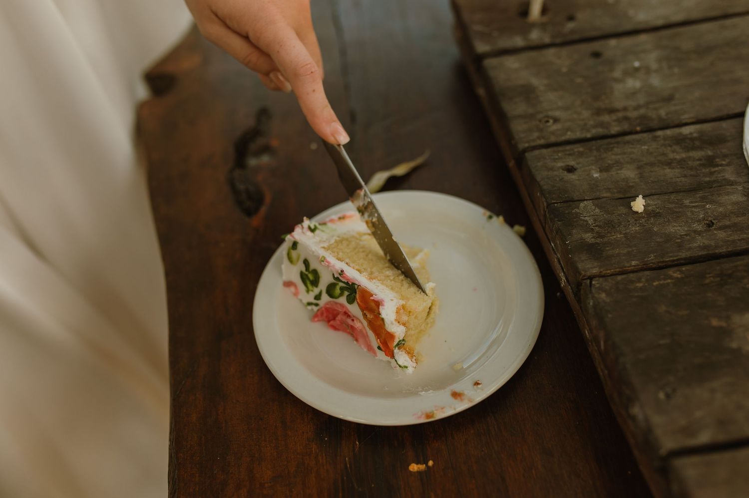 Hand cutting a slice of decorated wedding cake with a knife on a white plate on a wooden surface.