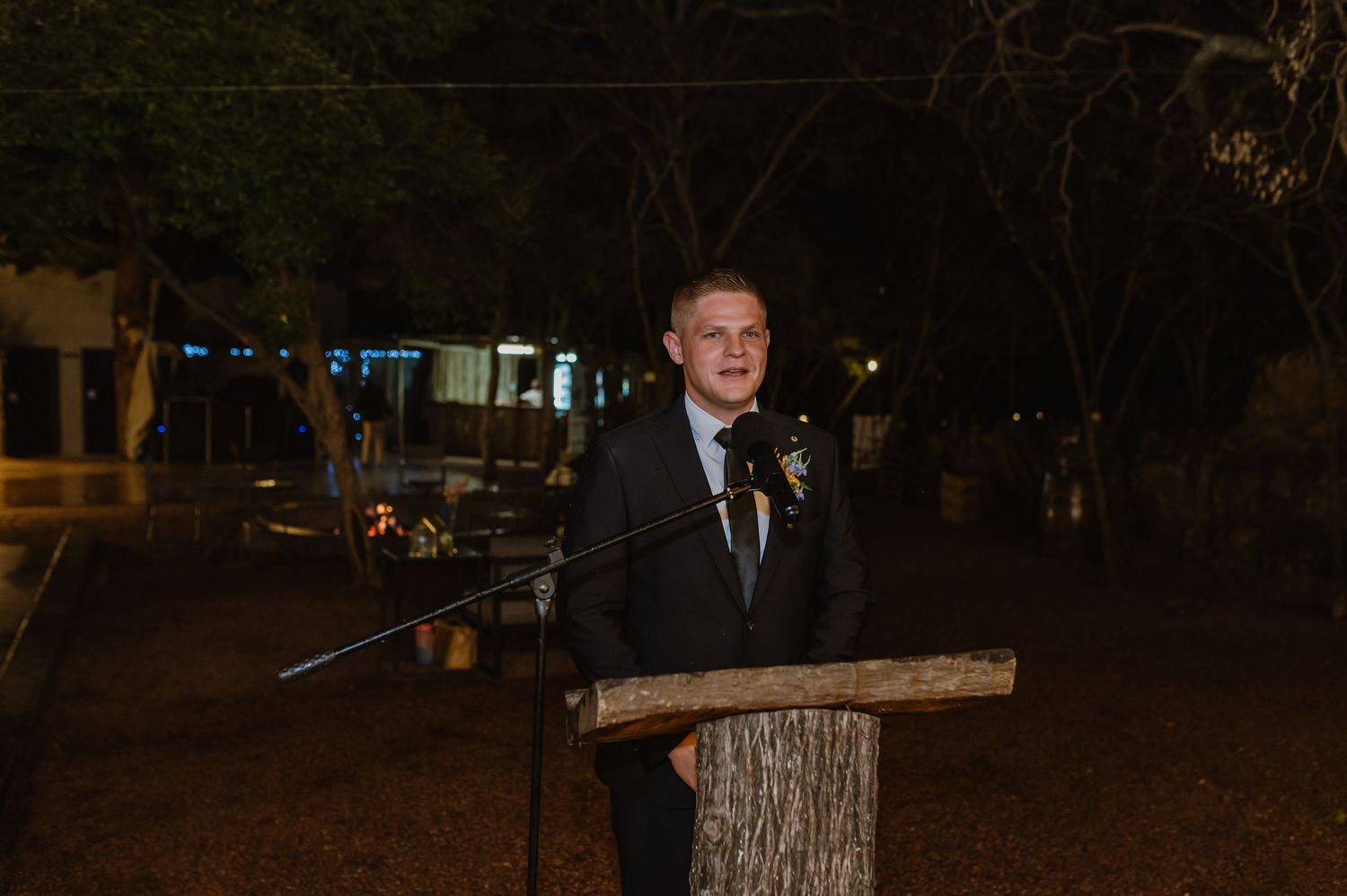 Groom in black suit delivering a speech at a rustic wooden podium at night.