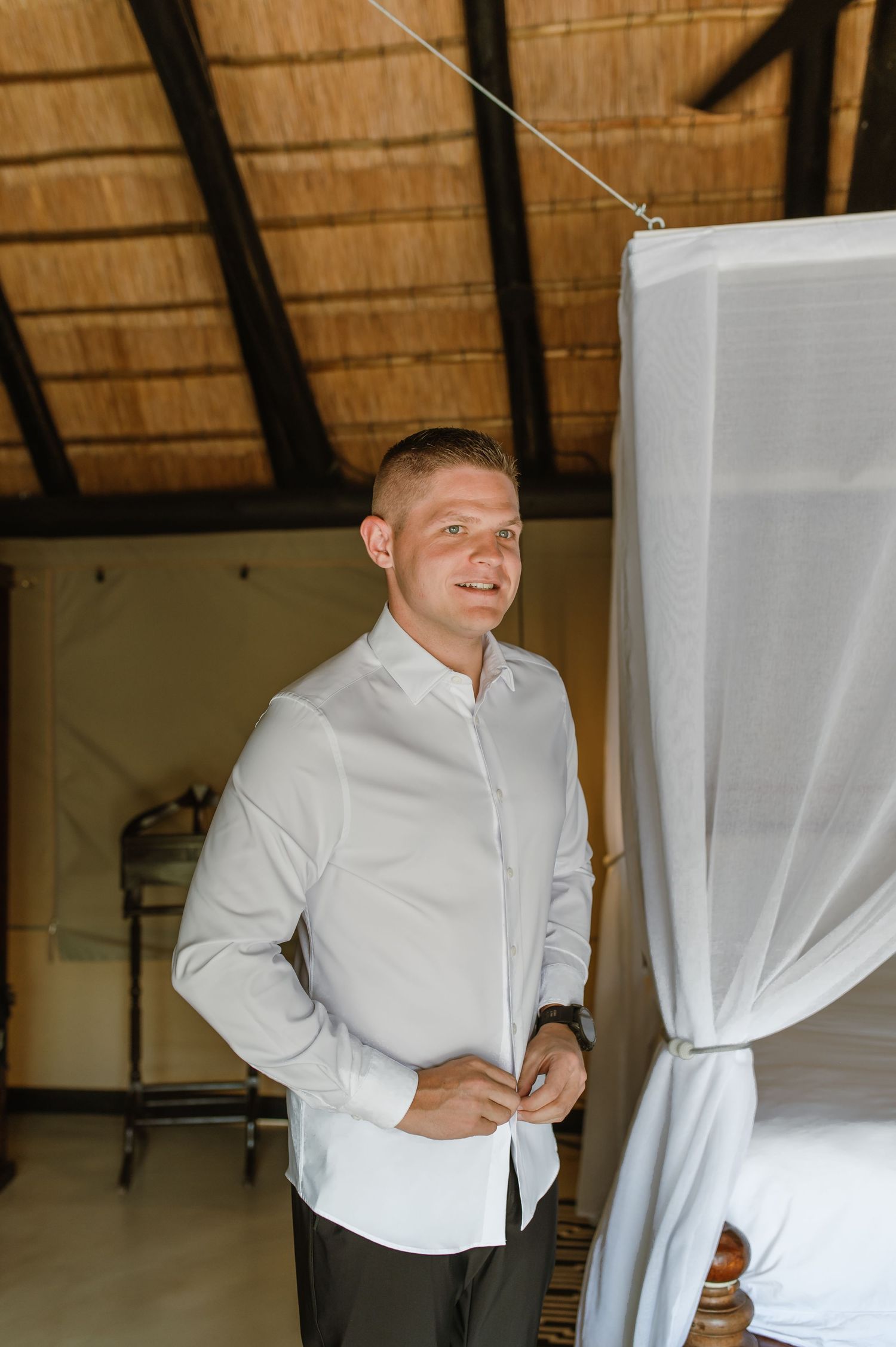 Smiling groom in a white shirt buttoning up inside a thatched-roof safari lodge room with a canopy bed.