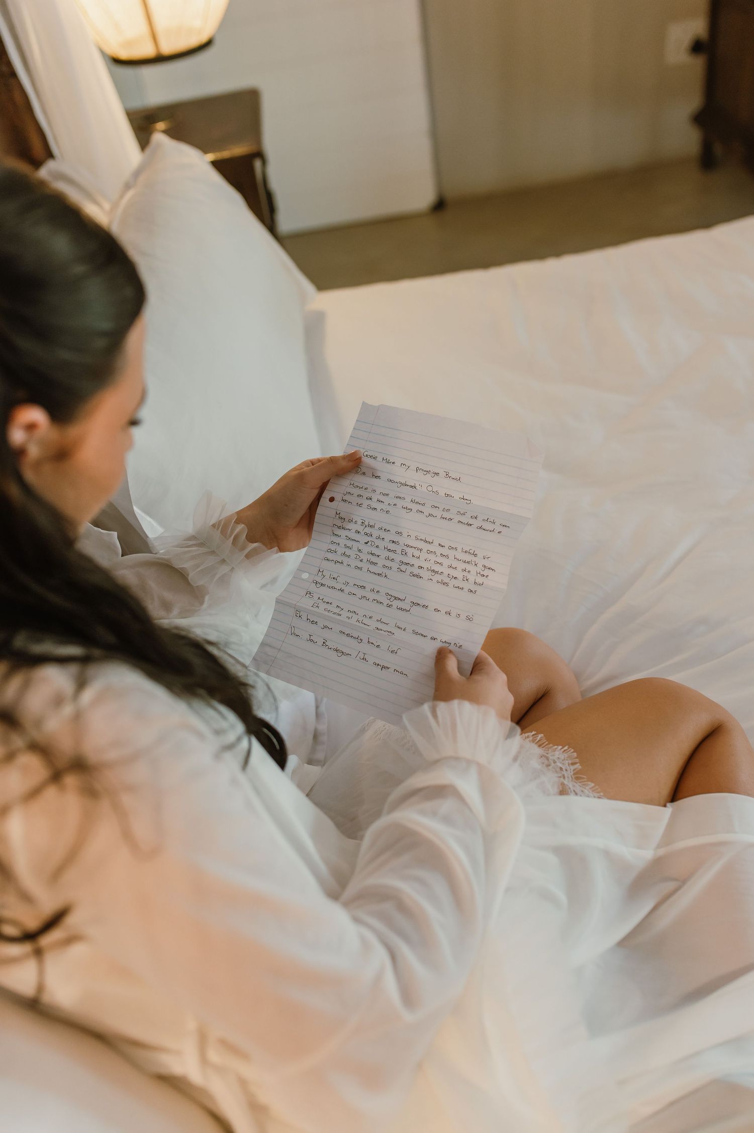 Bride in white robe reading a heartfelt handwritten letter while relaxing on a white bed.