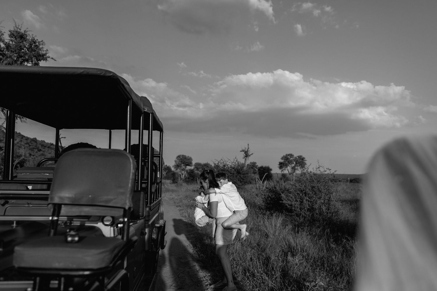 Black and white safari photo of a couple embracing beside a game drive vehicle in African bush landscape.