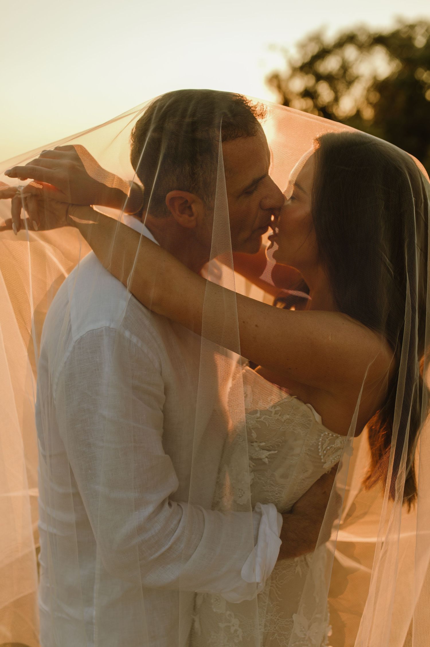 Bride and groom sharing romantic kiss under sheer veil at golden hour sunset wedding.