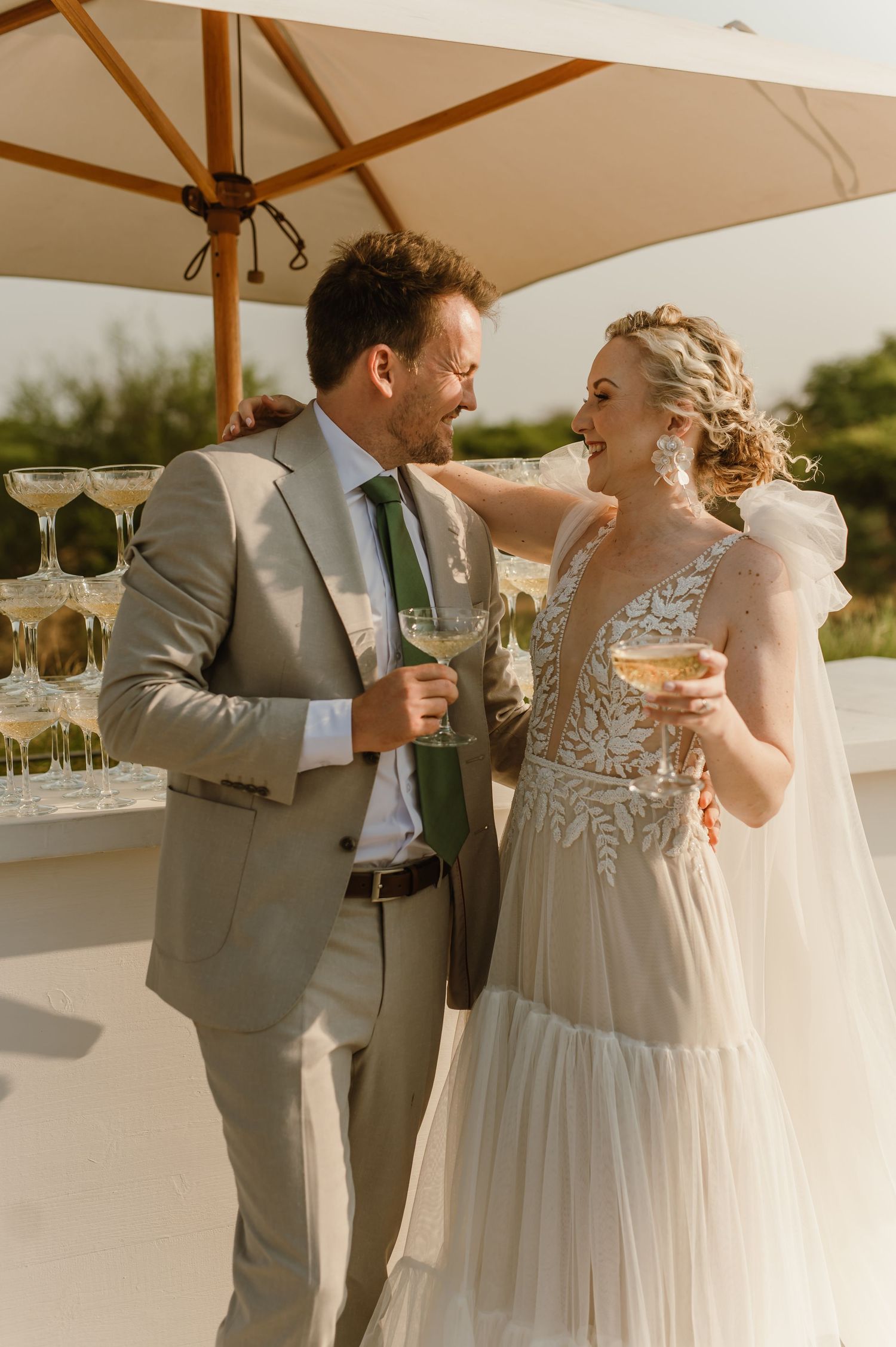 Couple shares intimate moment with champagne under outdoor umbrella during wedding reception.