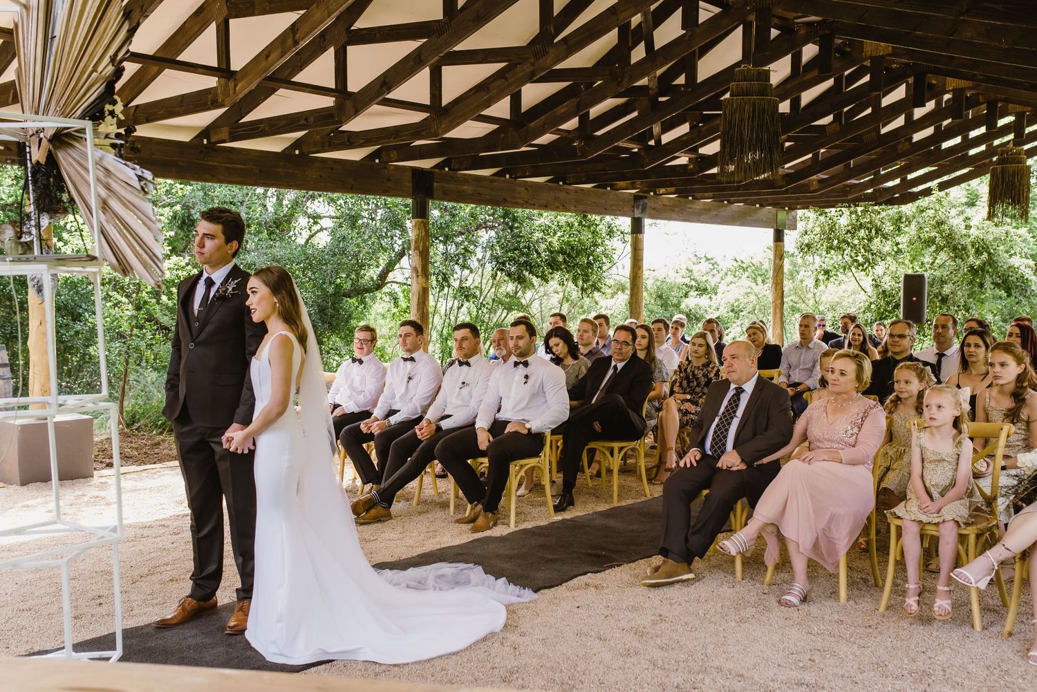 Wedding celebration under rustic pavilion with guests seated on wooden chairs and natural surroundings.