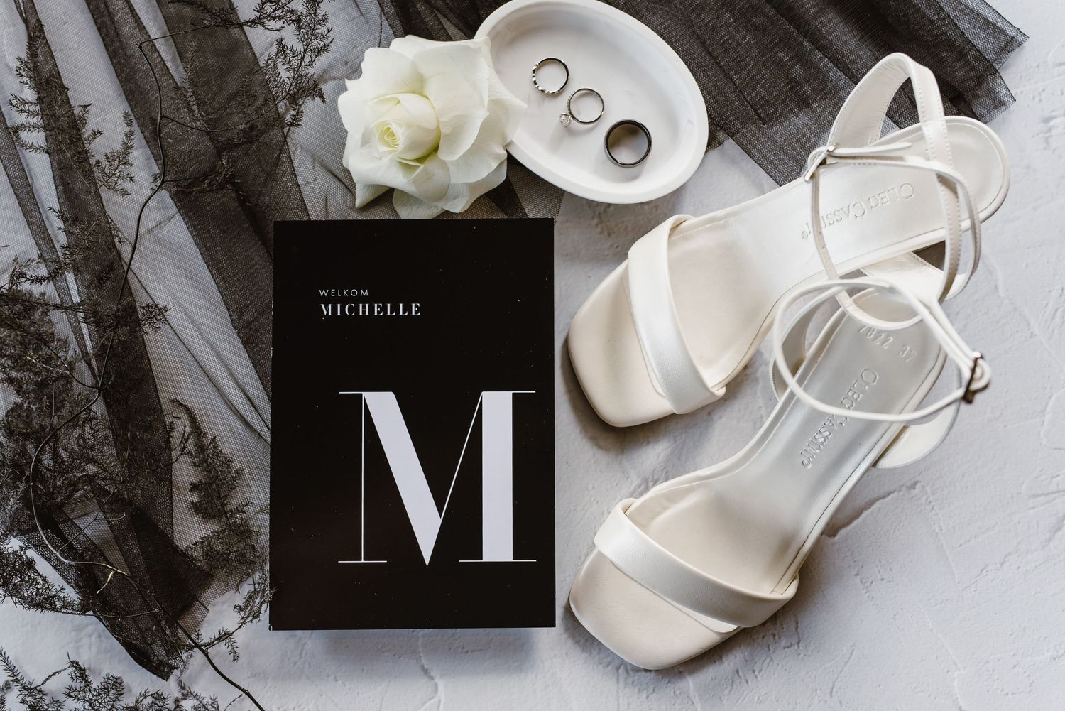 Wedding detail flatlay with rings, white roses, and elegant monogram on light background.