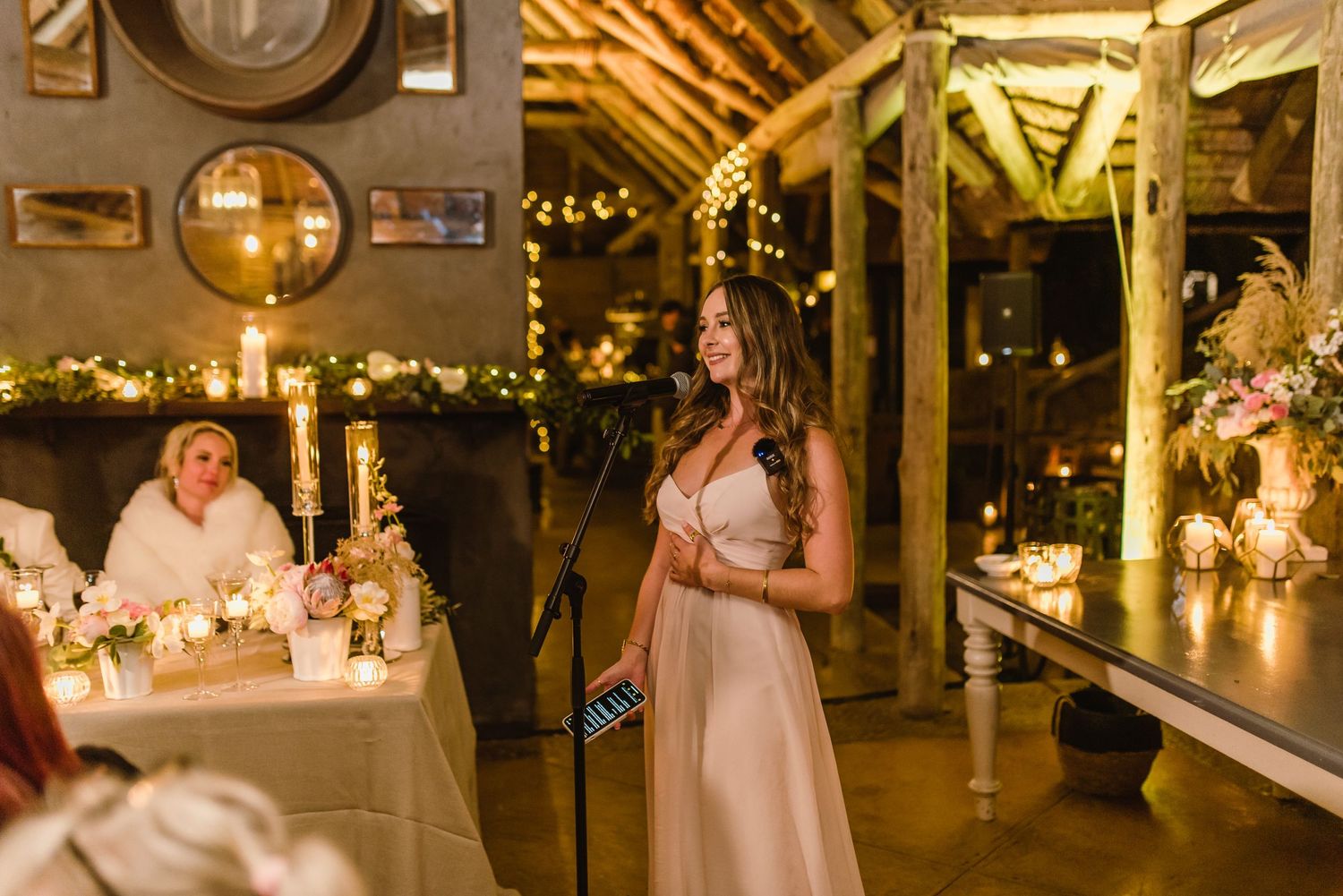 A speech being given at an elegant wedding reception with candlelit tables and rustic wooden beams overhead.