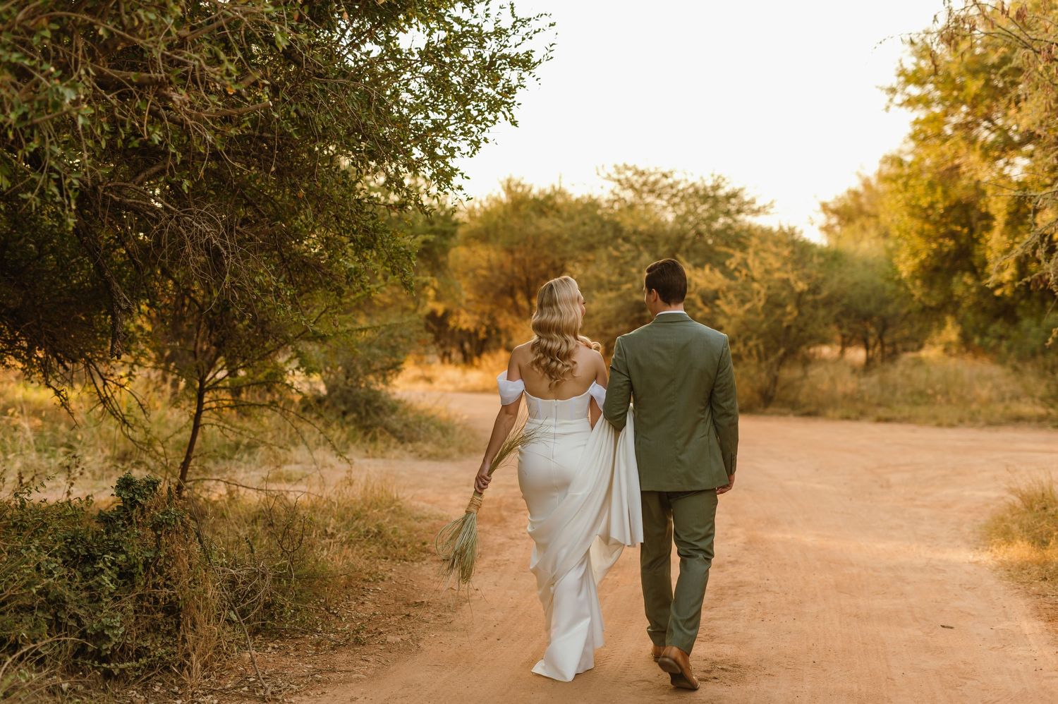 Couple walks together on a dirt path during golden hour surrounded by trees.
