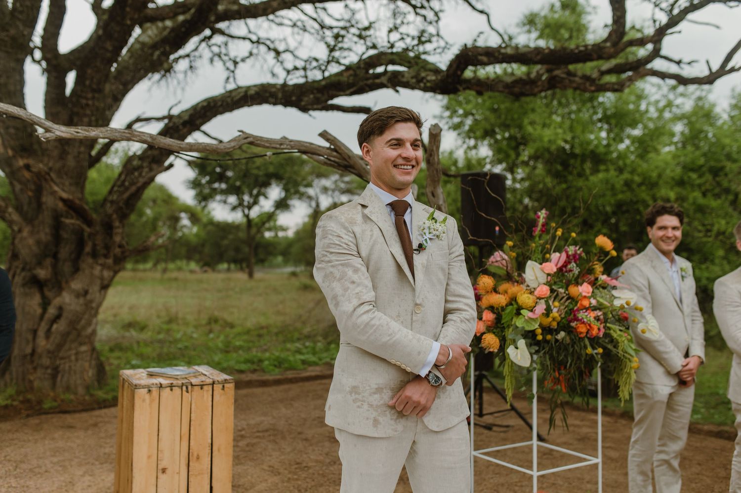 Wedding ceremony setup with floral arrangements and wooden elements under a large desert tree.