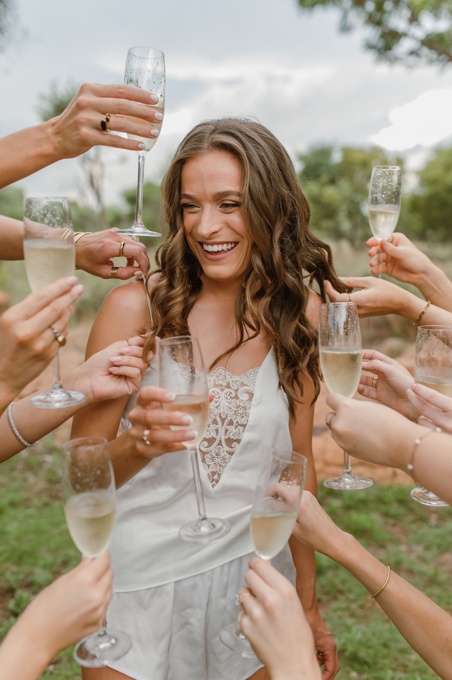 Woman in white lace dress celebrates with friends holding champagne glasses around her outdoors.