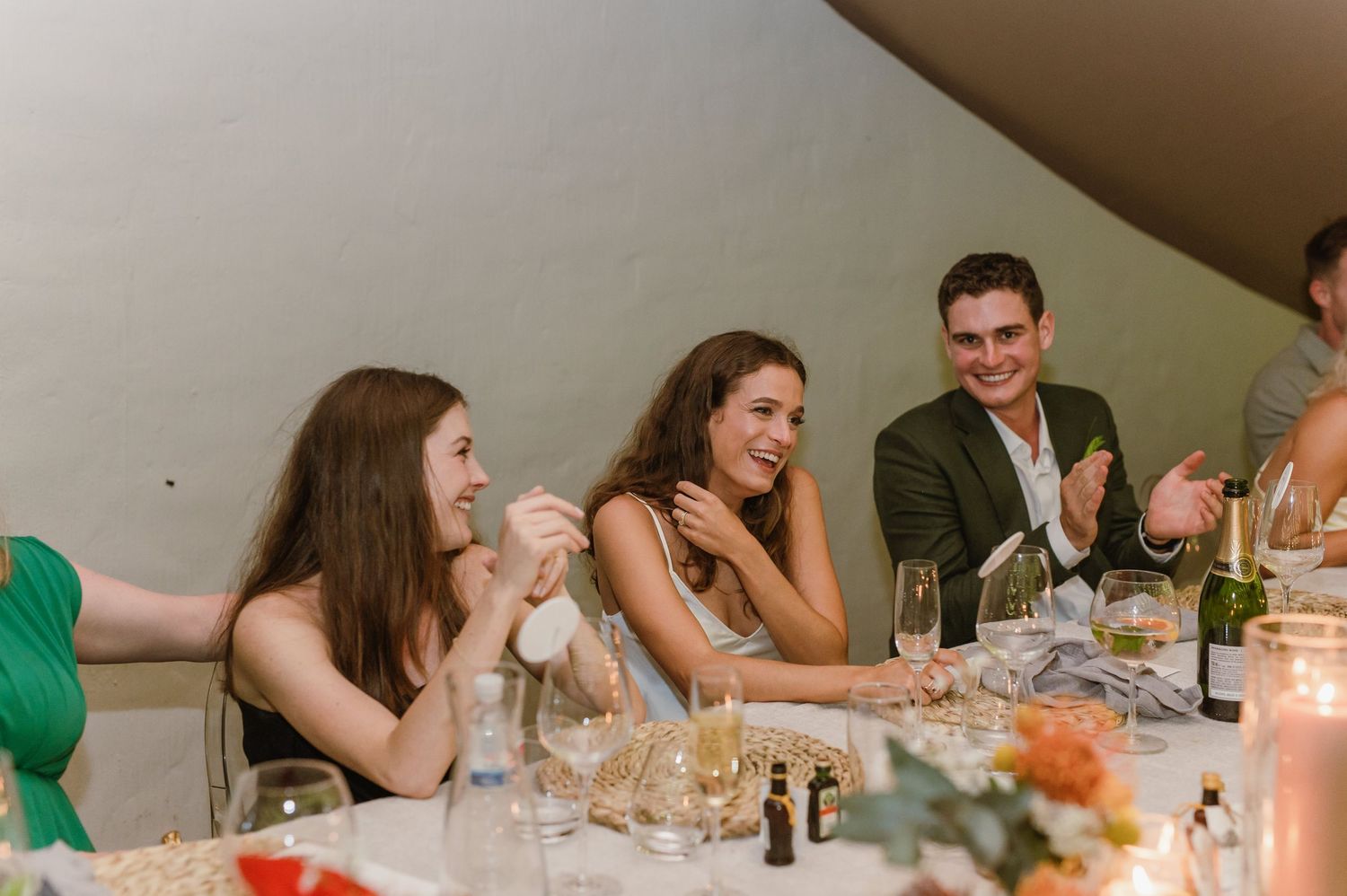 Wedding guests laugh and socialize at a reception dinner table with candlelight and floral centerpieces.
