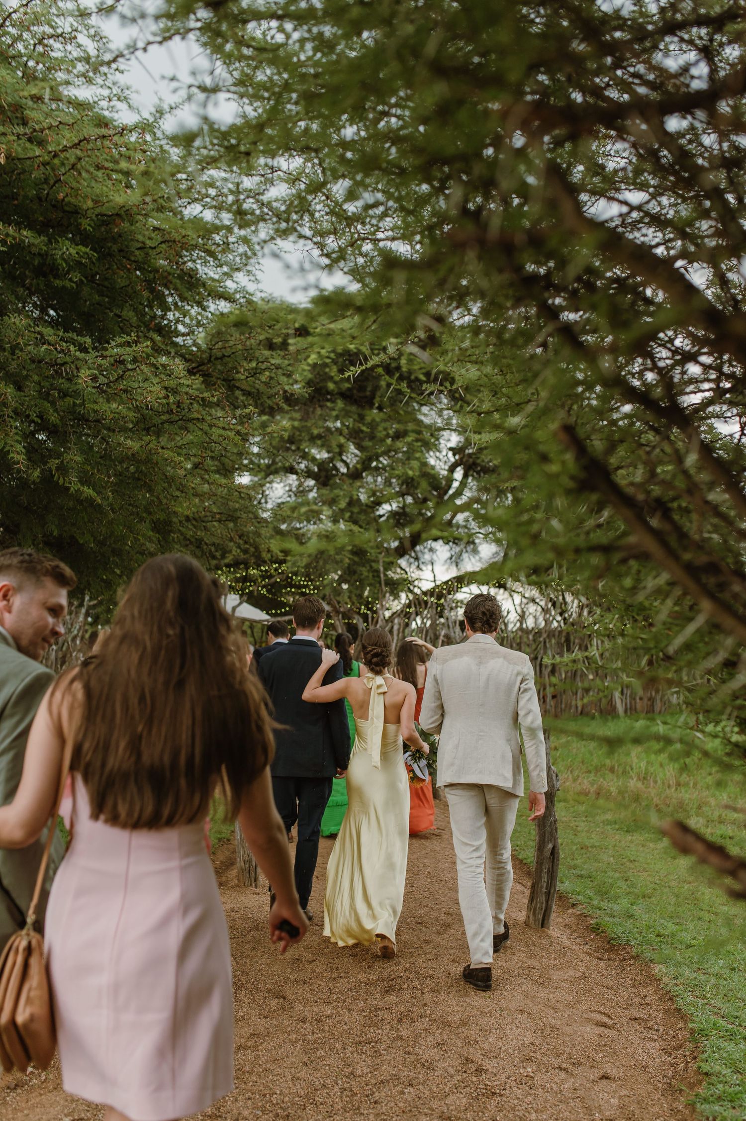 Wedding guests strolling down a tree-lined dirt path during an outdoor celebration.