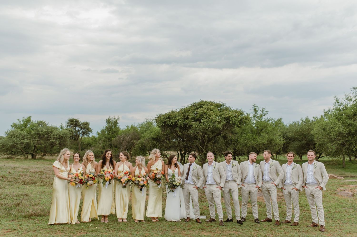Full wedding party in cream and beige attire posed for panoramic shot in natural setting.