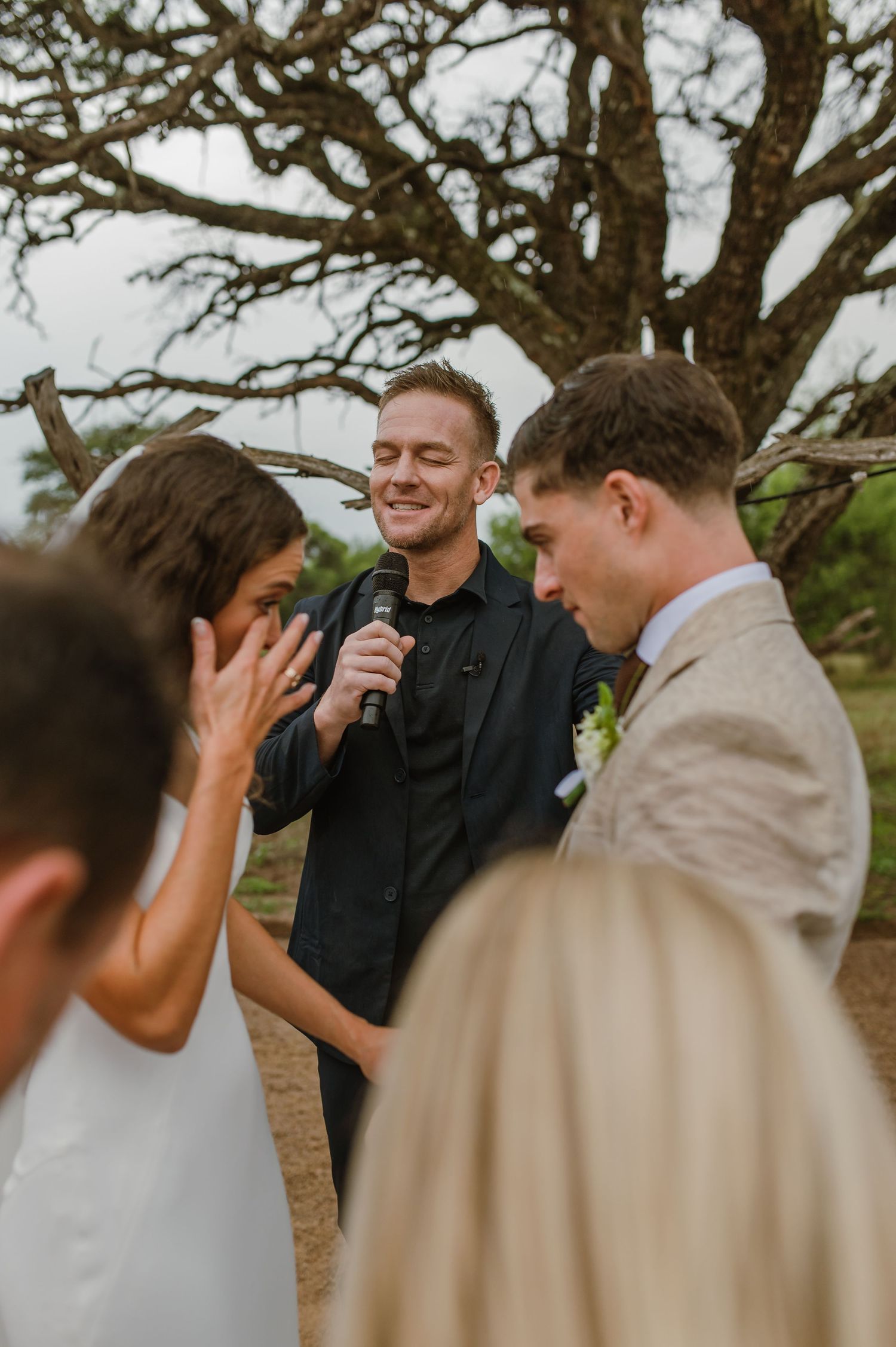 An emotional moment unfolds during an outdoor wedding ceremony beneath a sprawling tree.