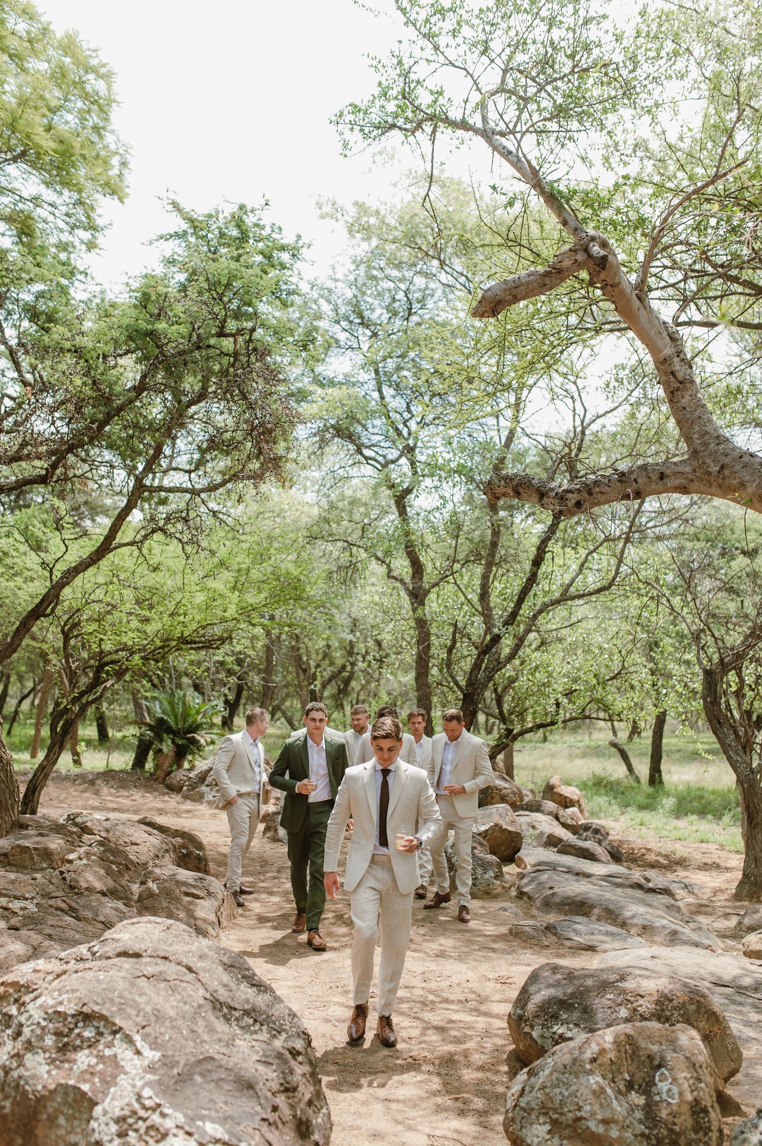 Wedding party in light suits gathered among rocky terrain and trees for casual photos.