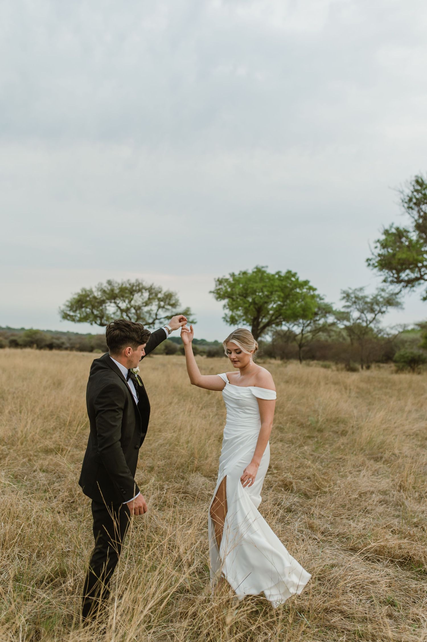 A couple in wedding attire dances and twirls together in a golden field at sunset.