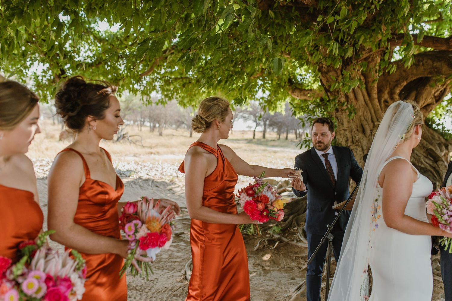 Wedding party stands in formation under shady trees during an outdoor ceremony.