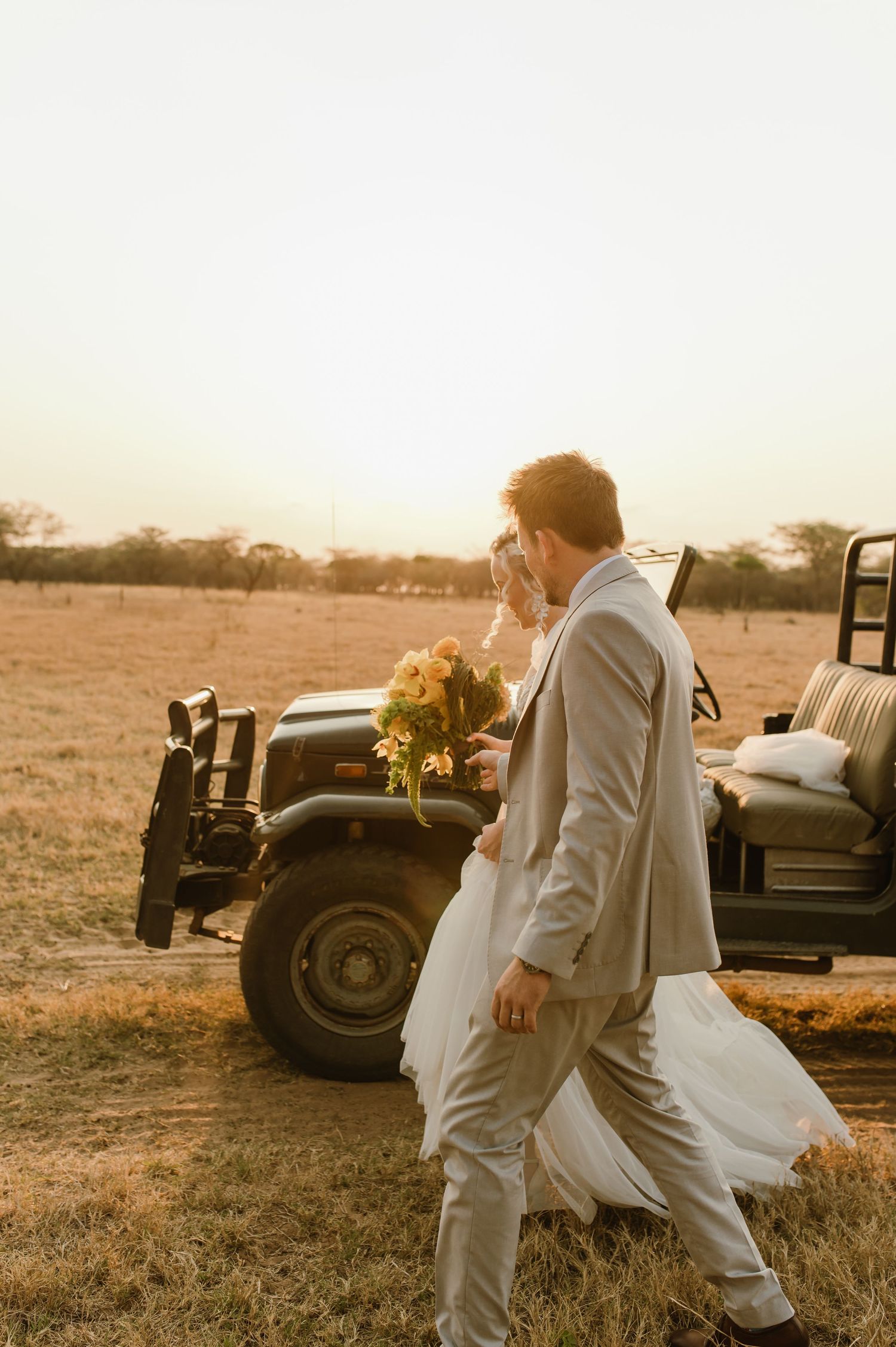 Newlyweds walking past classic vehicle in safari-style wedding setting at sunset.