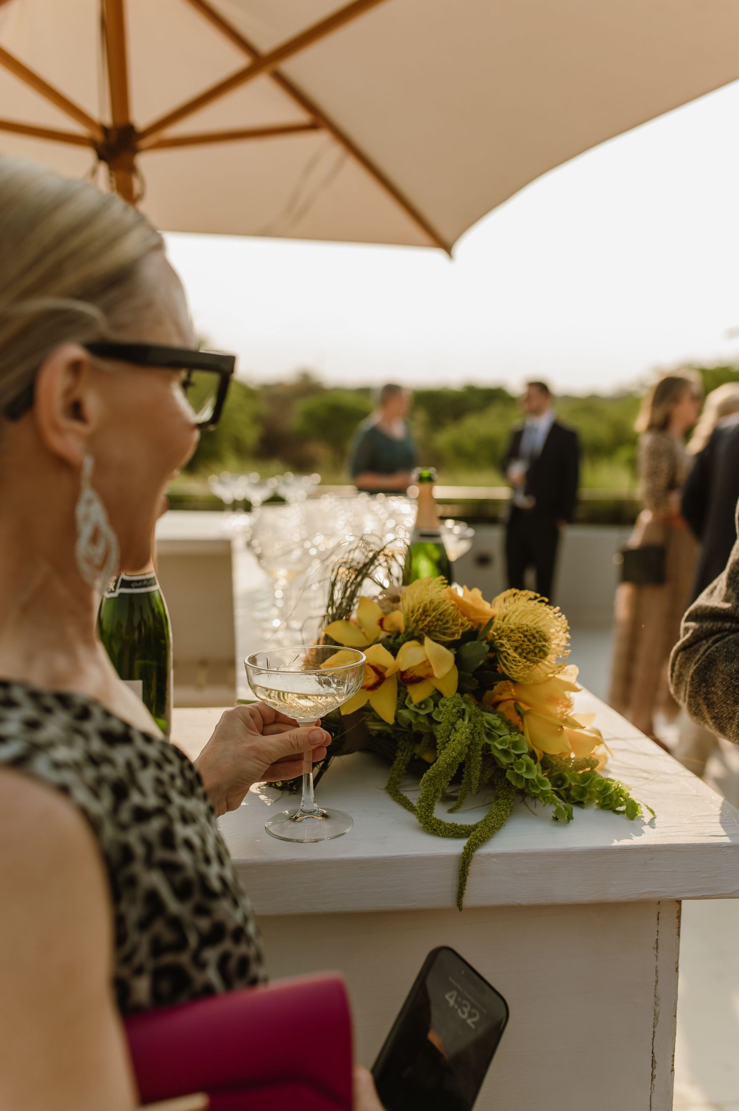 White table with yellow floral arrangements and glassware under beige patio umbrellas.
