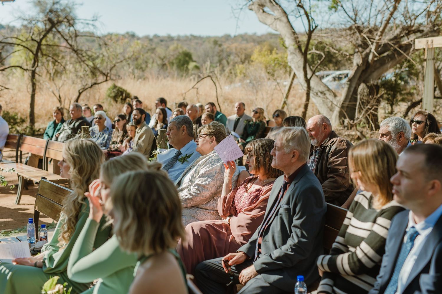 Wedding guests seated in wooden pews under natural desert tree canopy during ceremony.