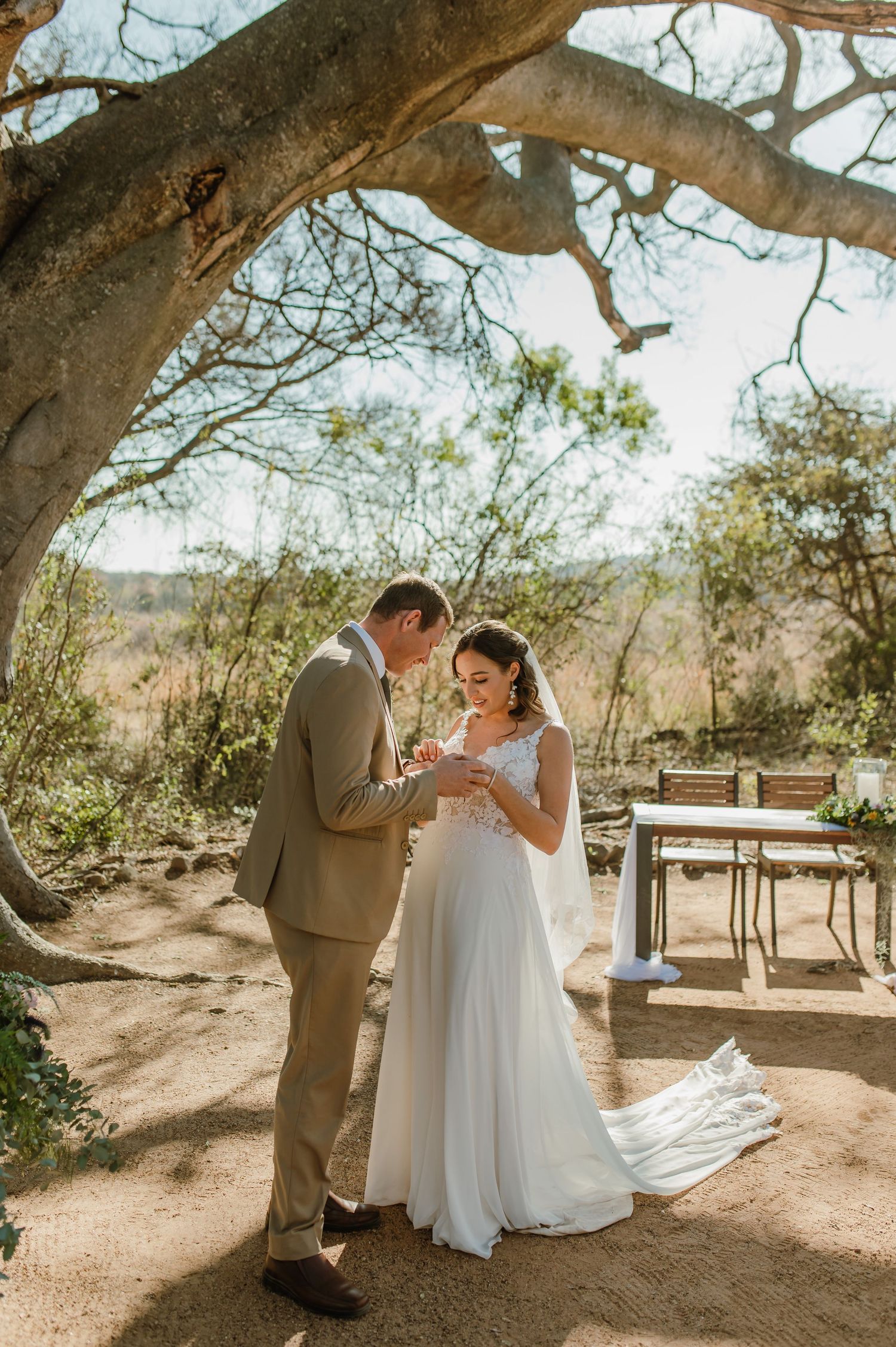 Newlyweds celebrate at wooden table beneath sprawling desert tree branches.