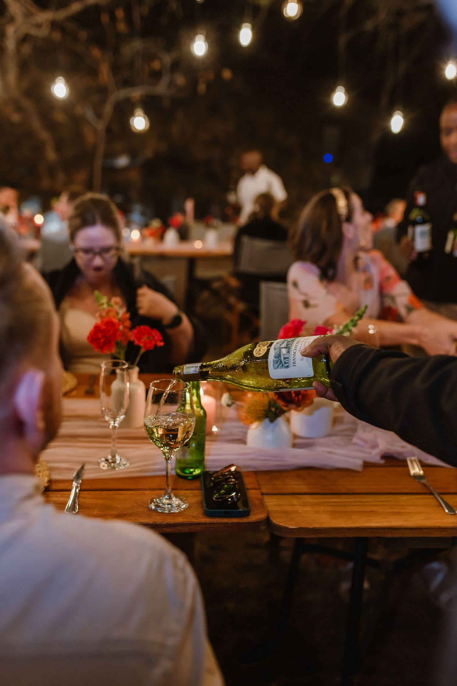 Wine glasses and table settings on rustic wooden tables under warm string lights at night.