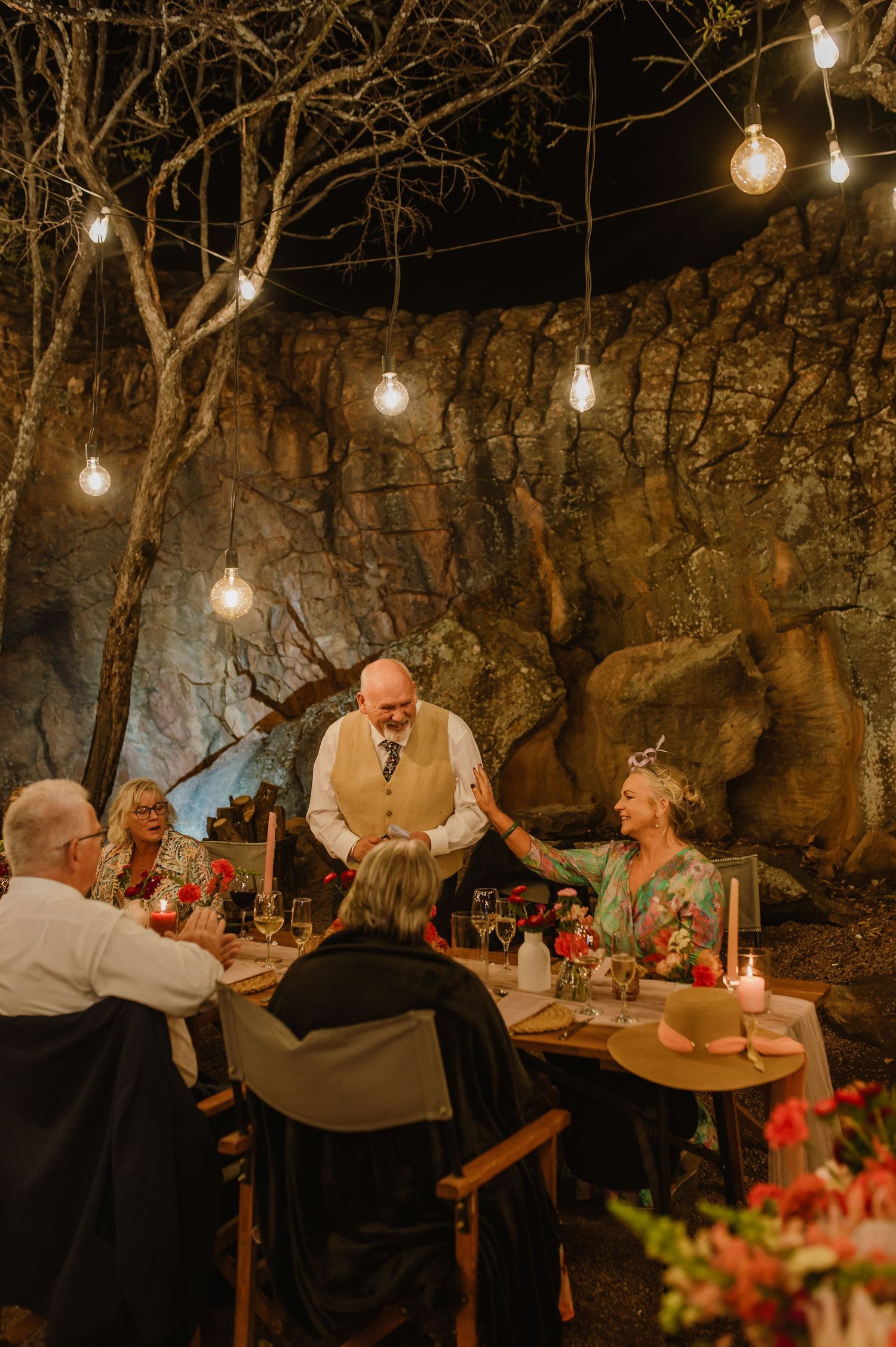 Evening dinner party with guests seated at long tables decorated with red flowers and candles in a cave setting.