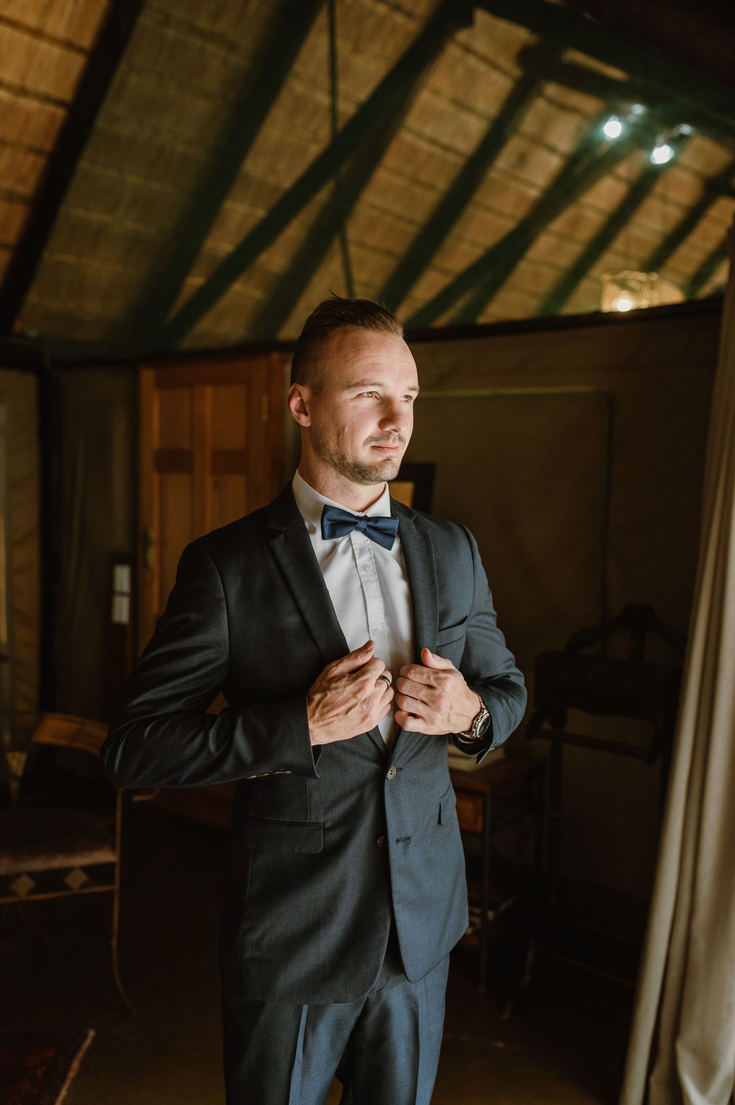 Person adjusting formal suit and tie in dimly lit room with wooden ceiling.