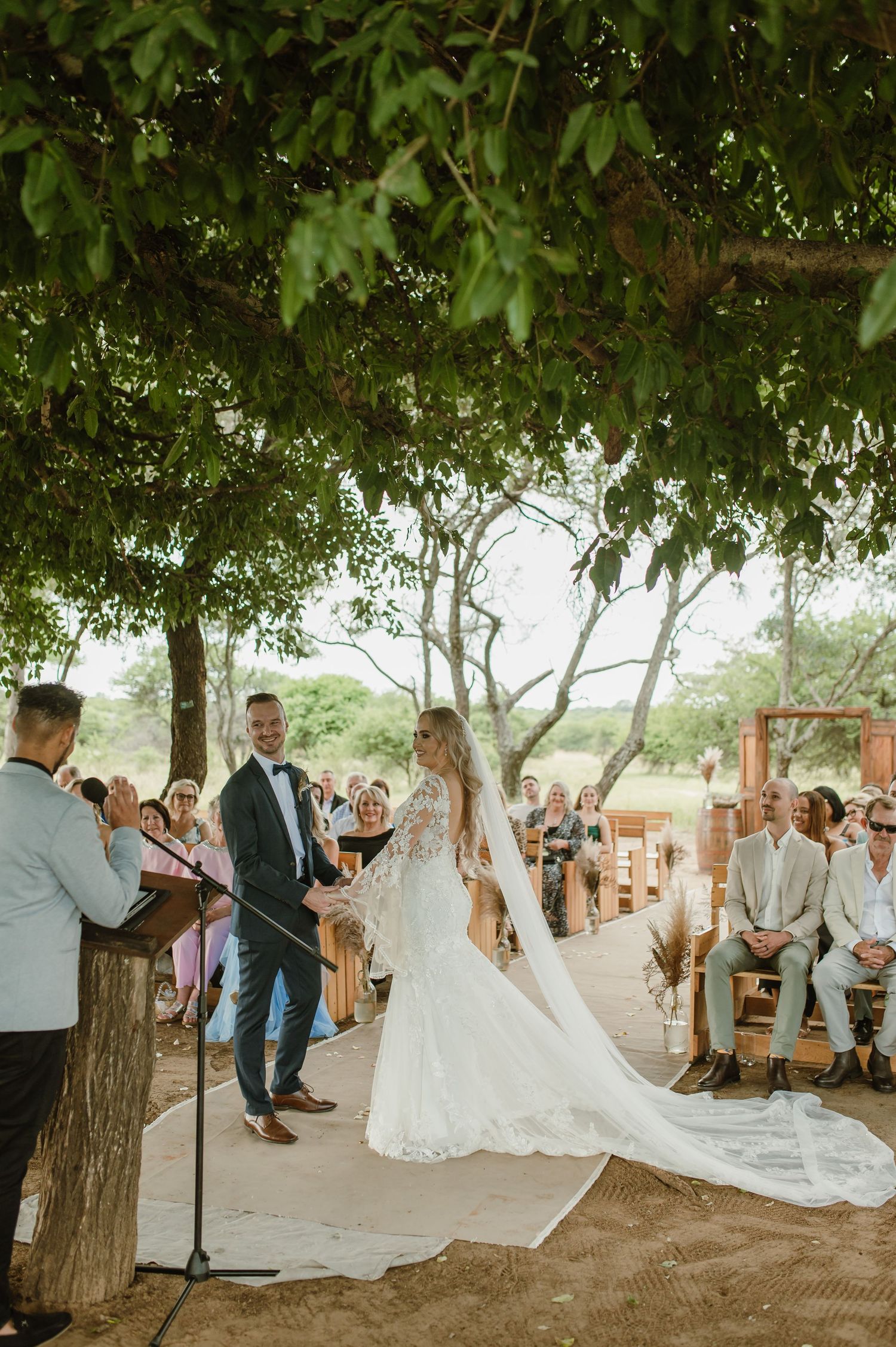 Wedding ceremony procession captured in panoramic view under leafy green trees.
