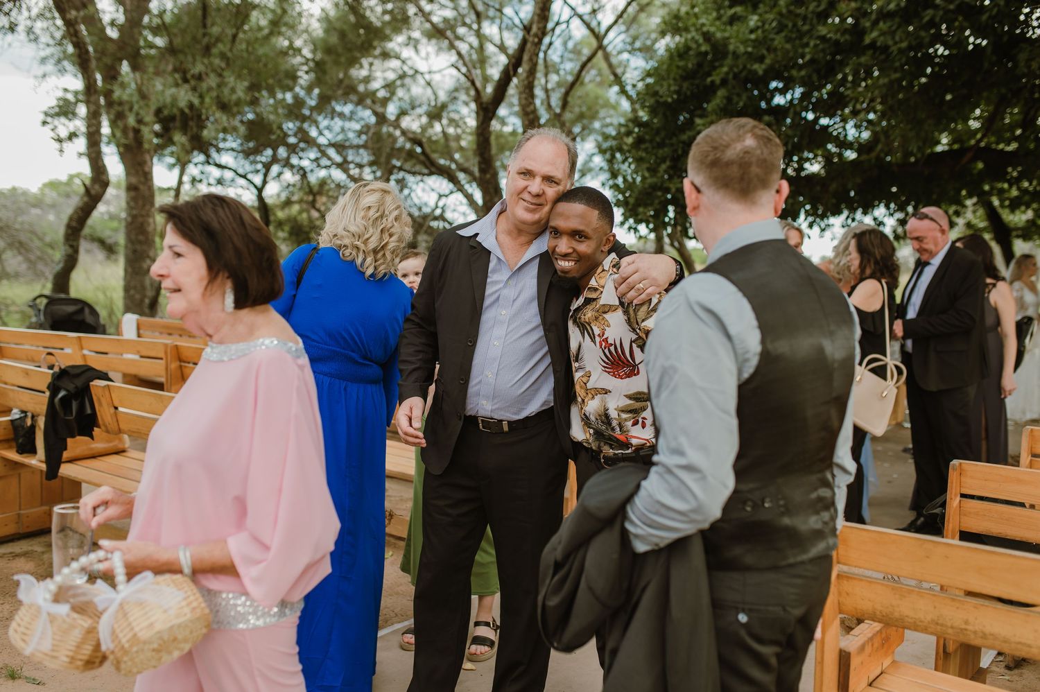 Wedding guests mingle and socialize between wooden benches at an outdoor reception.