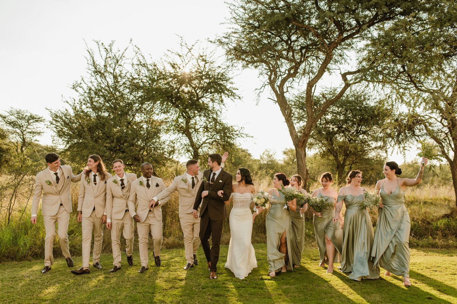 Joyful wedding party in coordinated sage and tan attire poses playfully outdoors.