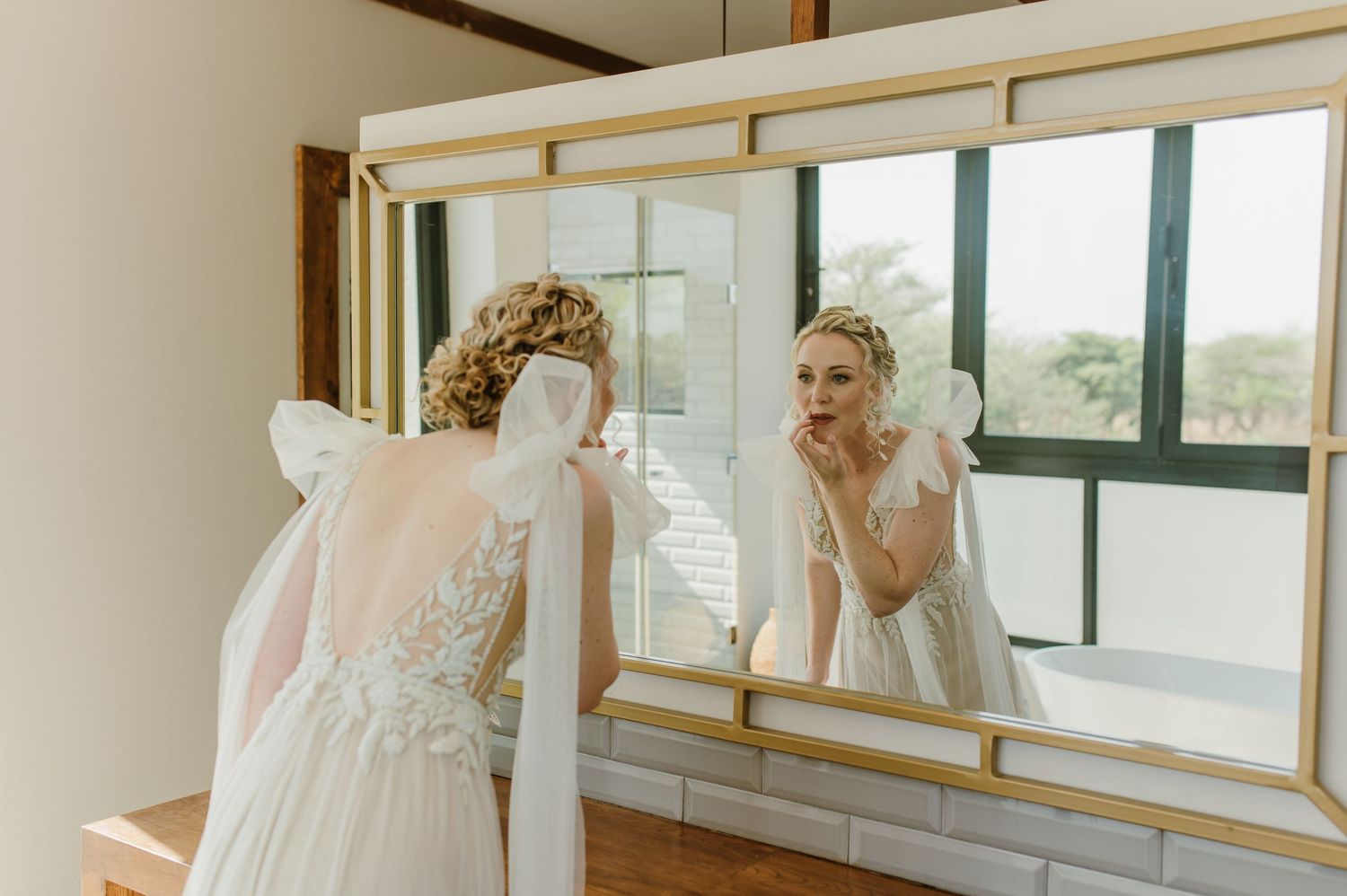 A bride stands in an elegant room with floor-to-ceiling windows and gold-framed mirrors.