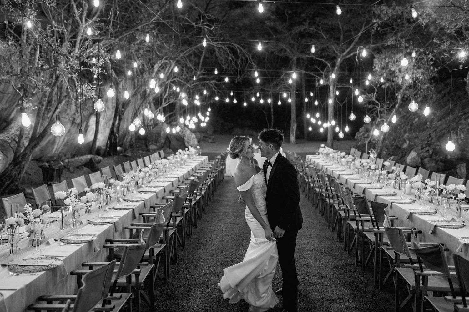 Black and white photo of outdoor wedding reception with string lights creating magical ambiance.