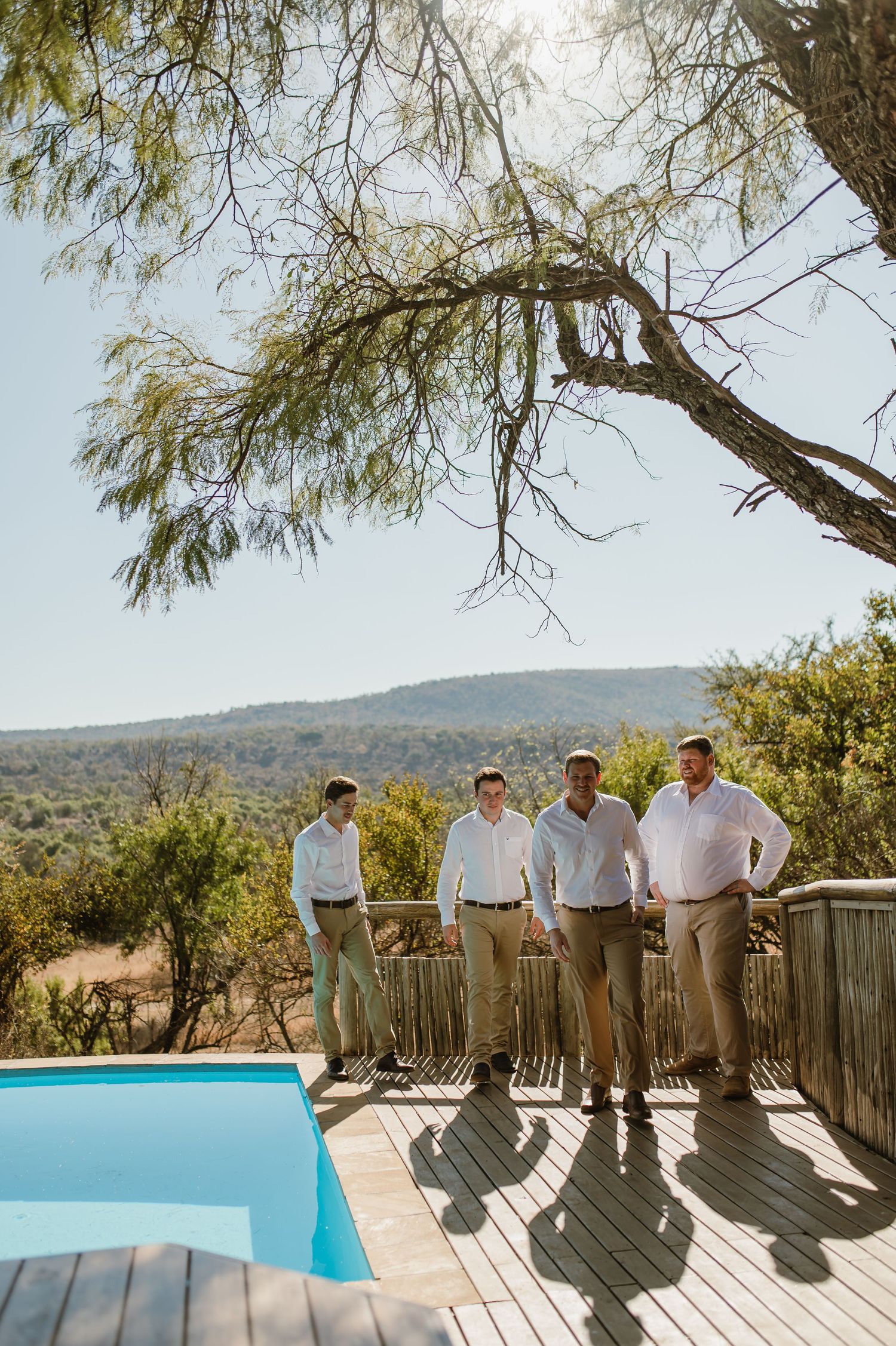 Wedding party members posed on wooden deck with swimming pool and mountain landscape.