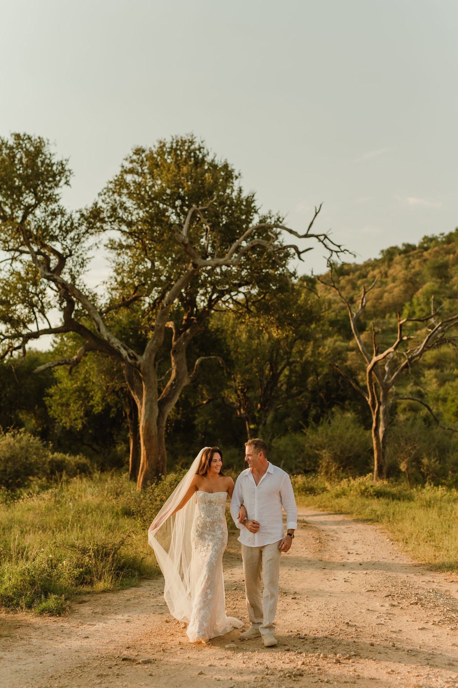 Bride and groom walk hand-in-hand down a sunlit dirt path surrounded by lush trees and rolling hills at golden hour.