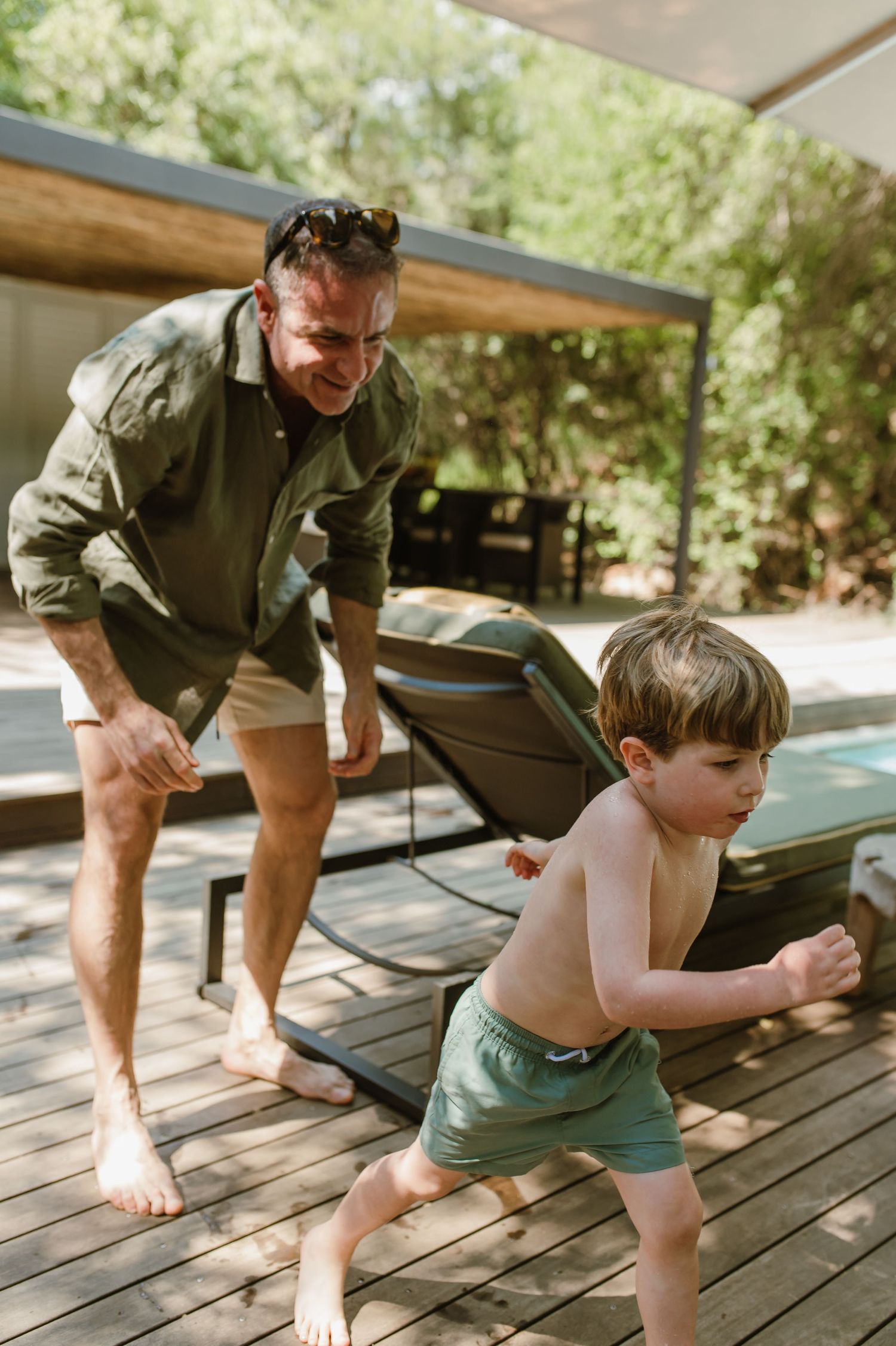 A man chases a young boy in green swim shorts on a wooden deck surrounded by lush trees.