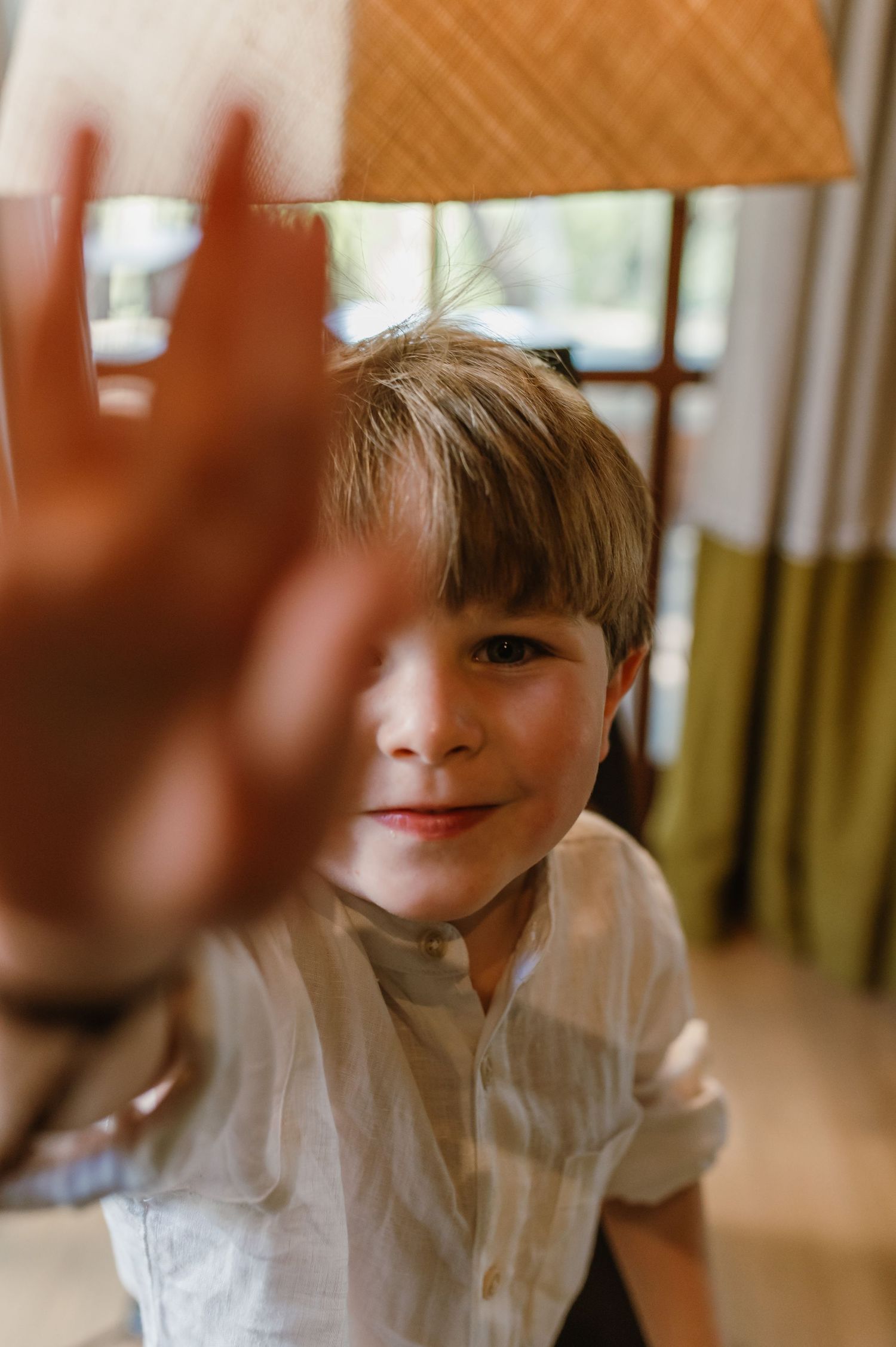 Young boy in white linen shirt playfully reaching toward camera with a mischievous smile indoors.