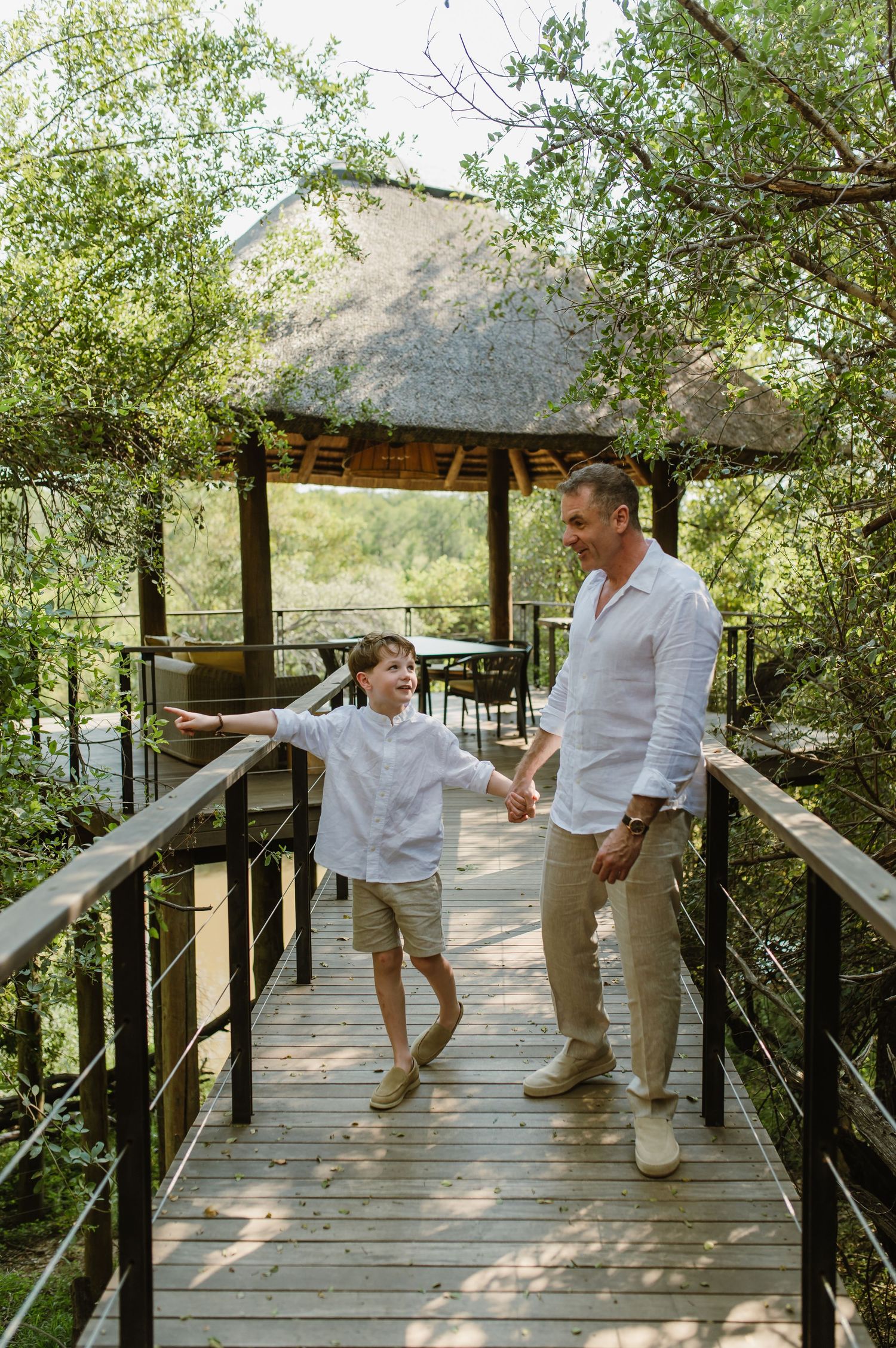 Father and son holding hands on wooden walkway surrounded by lush green trees at safari lodge.