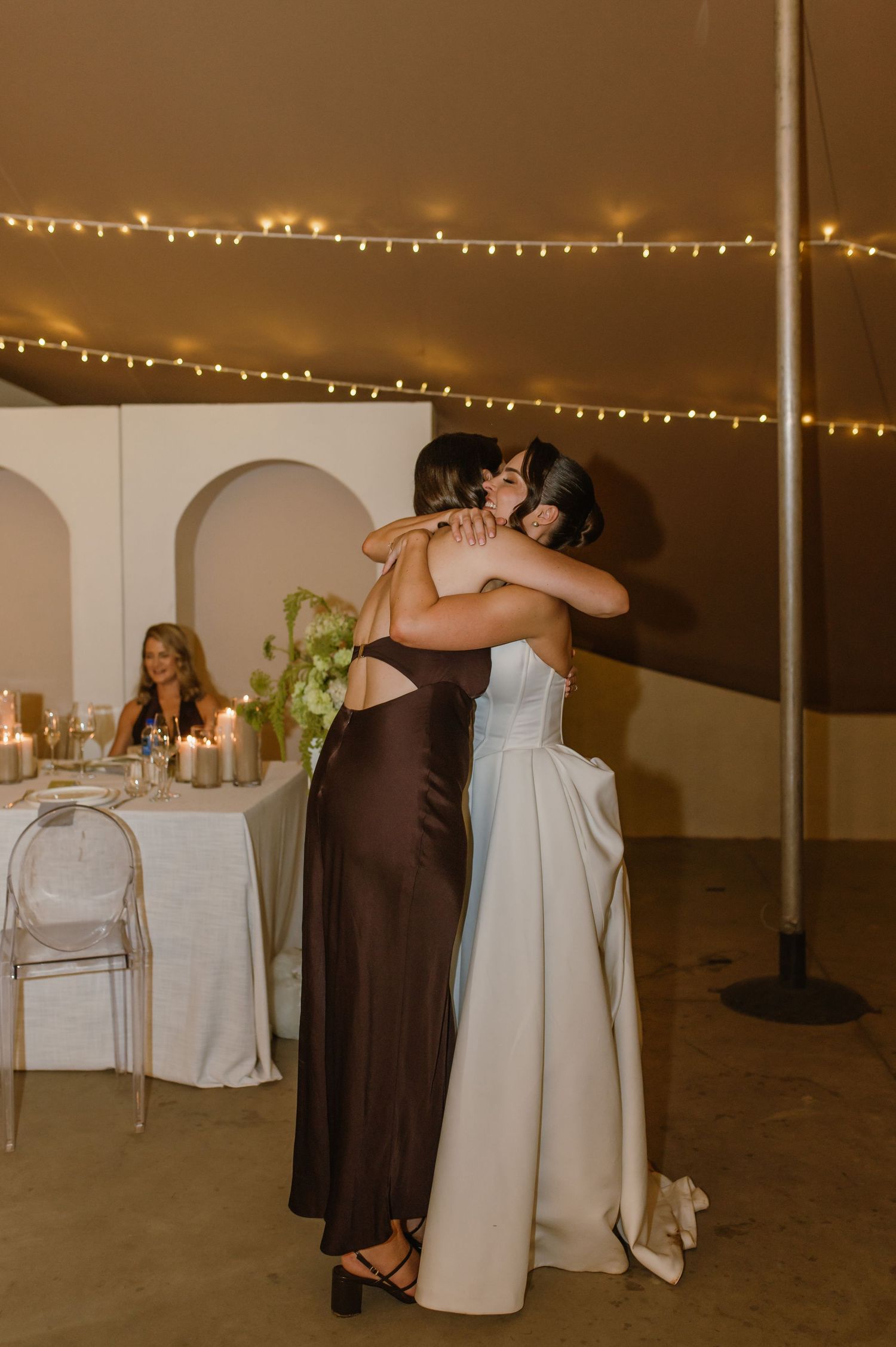 Two people share an emotional embrace at a wedding reception under warm string lights.