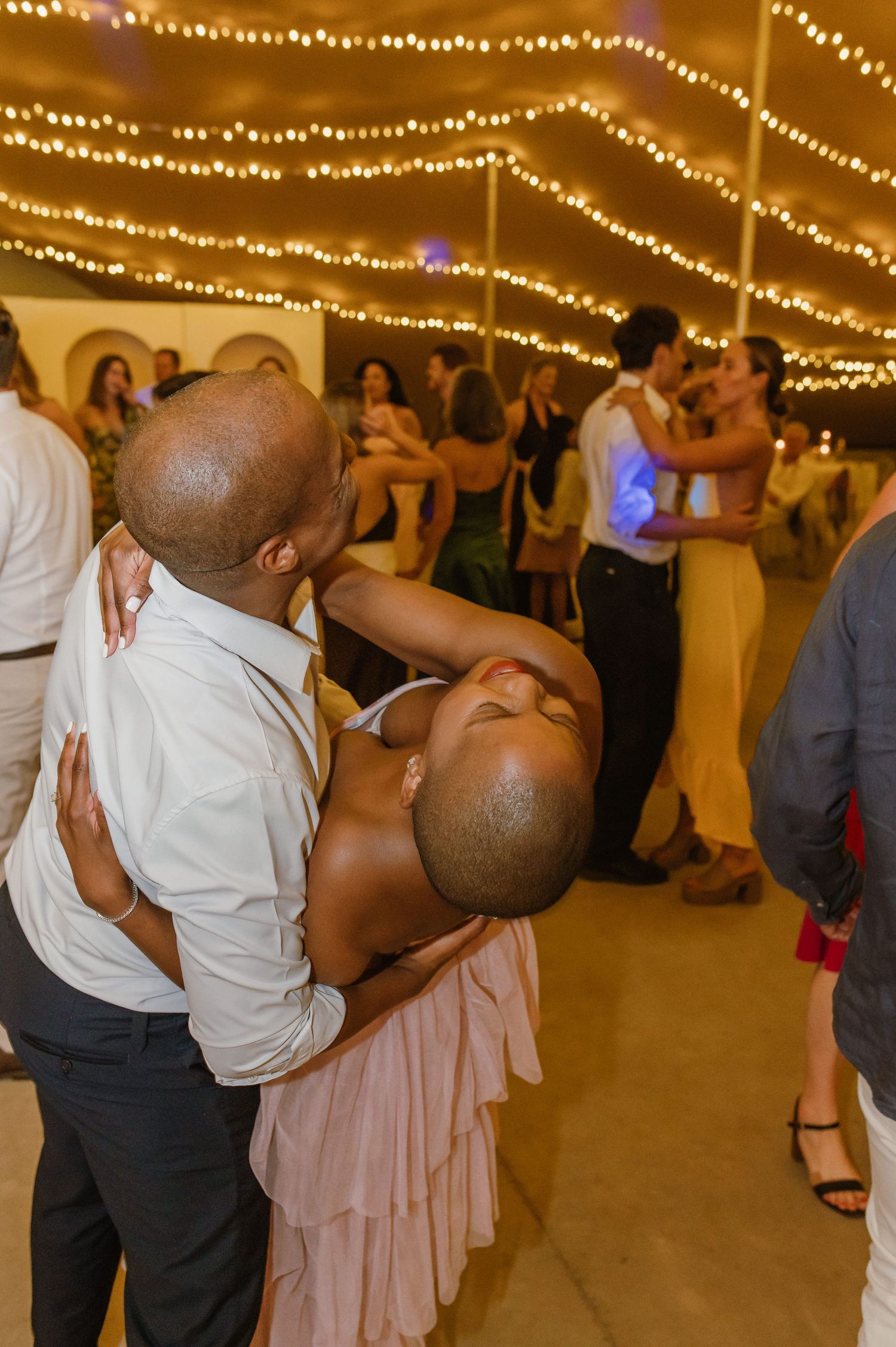 Wedding guests dancing and celebrating under warm string lights at an evening reception.