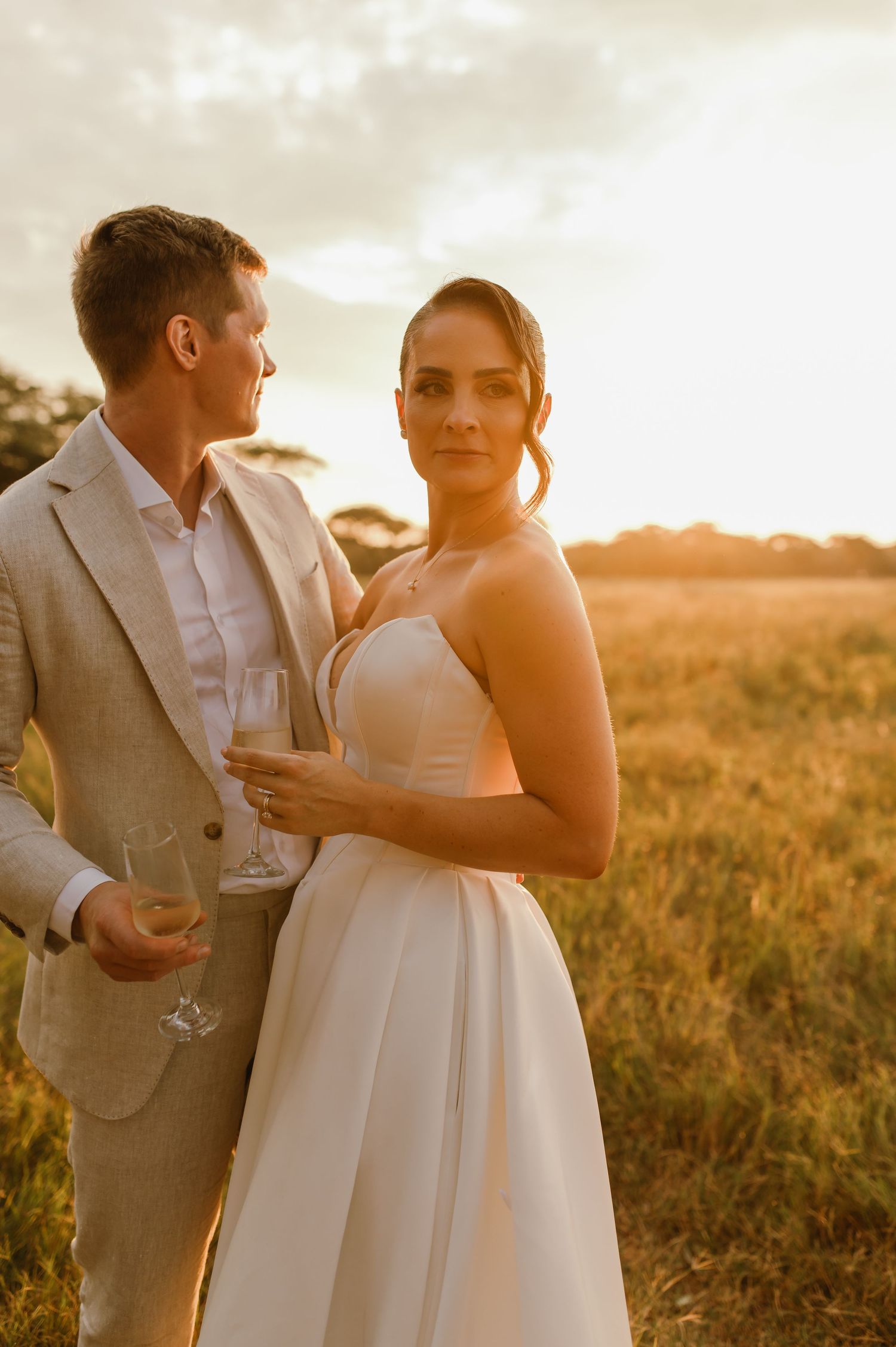 A tender moment between newlyweds sharing drinks in a sunlit savanna landscape.